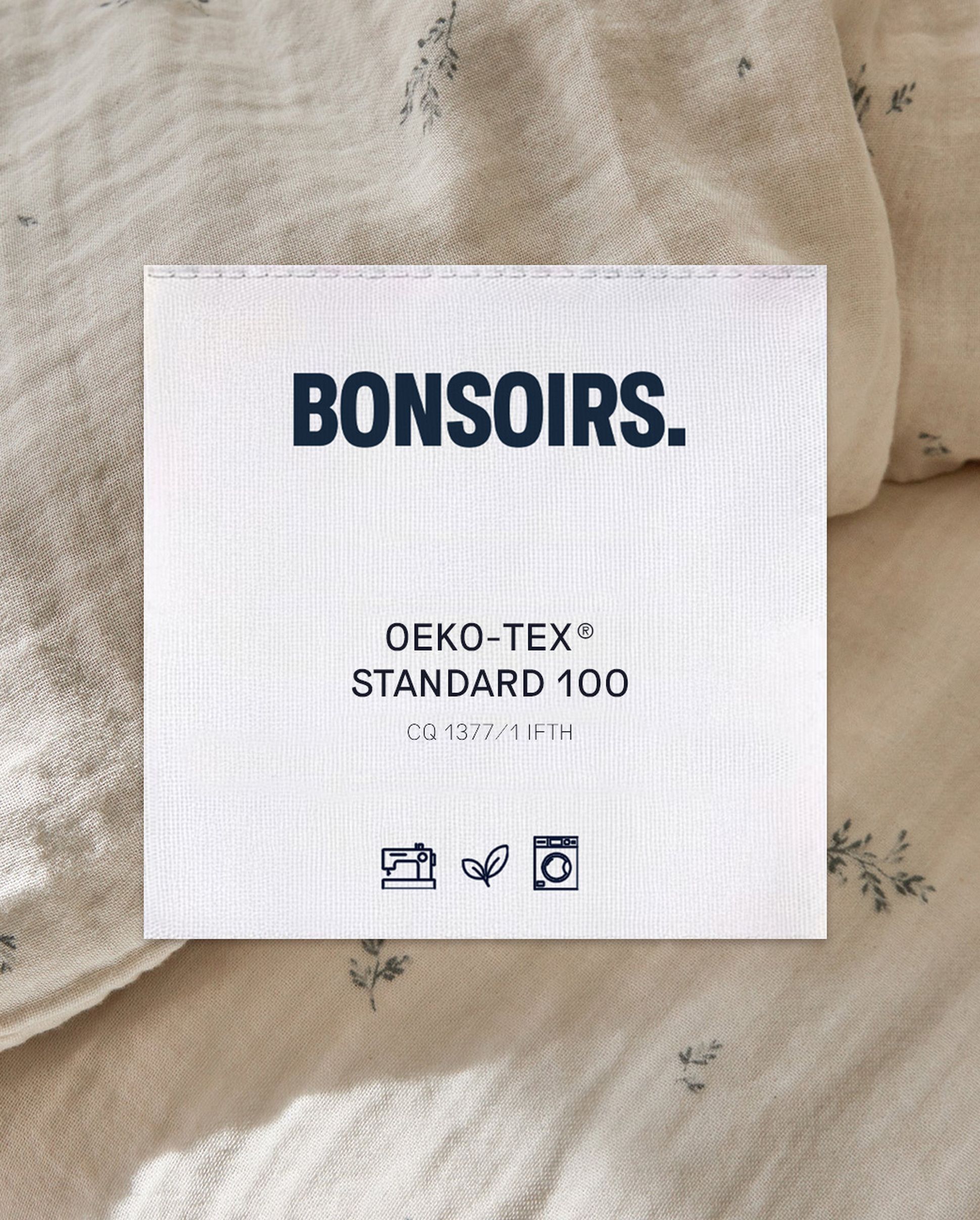 Une étiquette en tissu beige portant l'inscription « BONSOIRS » en caractères gras. Elle mentionne la certification « OEKO-TEX® STANDARD 100 » et comporte des symboles indiquant les consignes de lavage, de repassage et le respect de l'environnement.