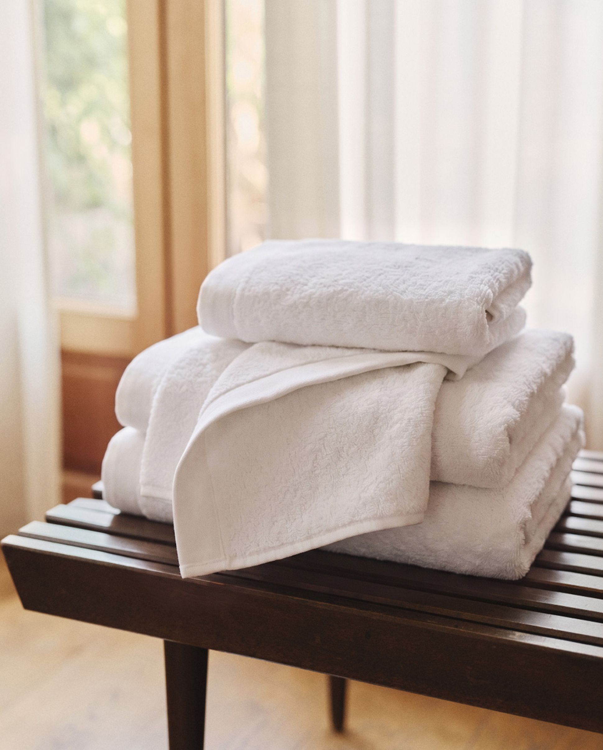 A stack of fluffy white towels neatly arranged on a wooden bench, with soft natural light filtering through sheer curtains in the background.