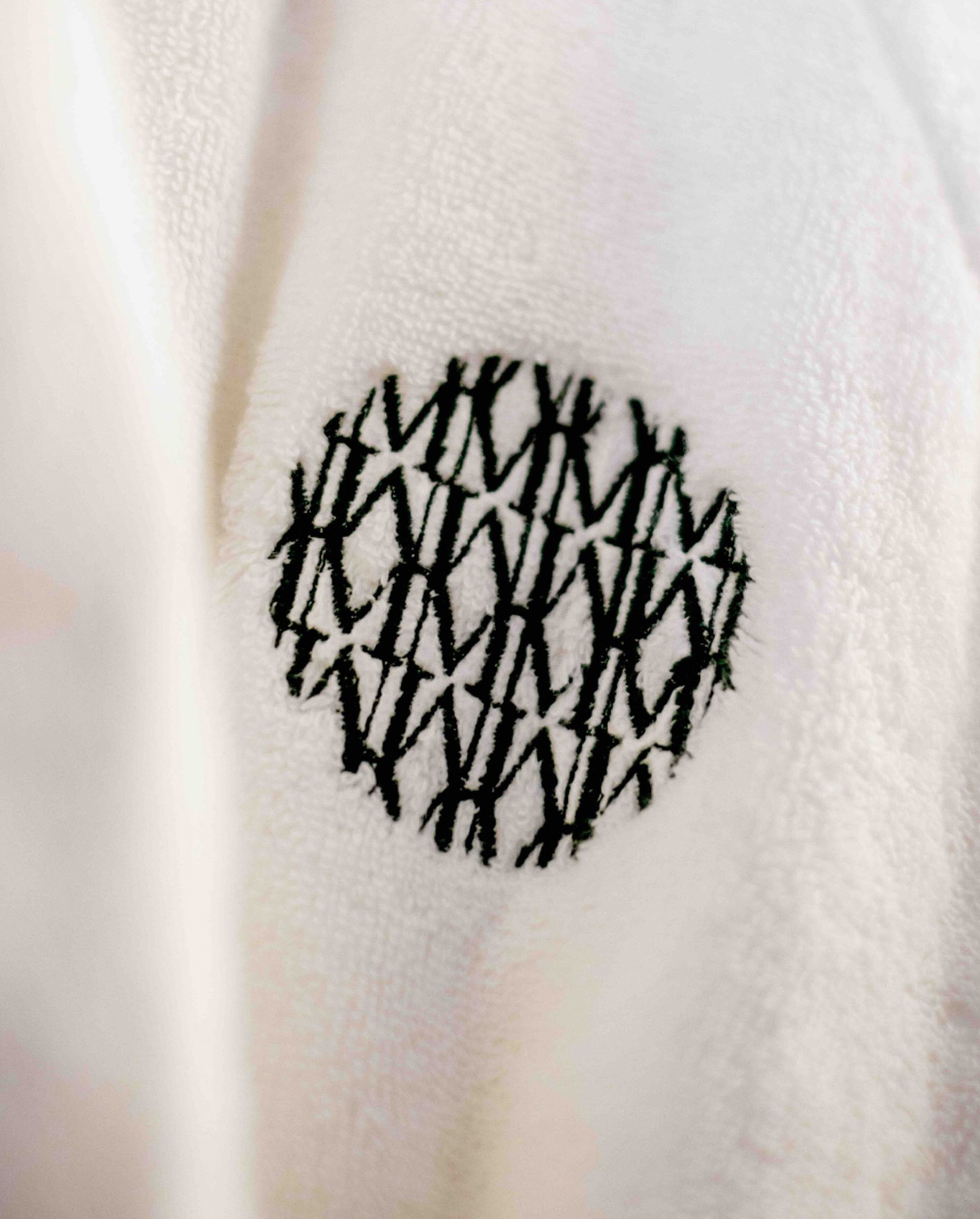 White fabric with a circular black embroidered pattern of overlapping letter "M"s.