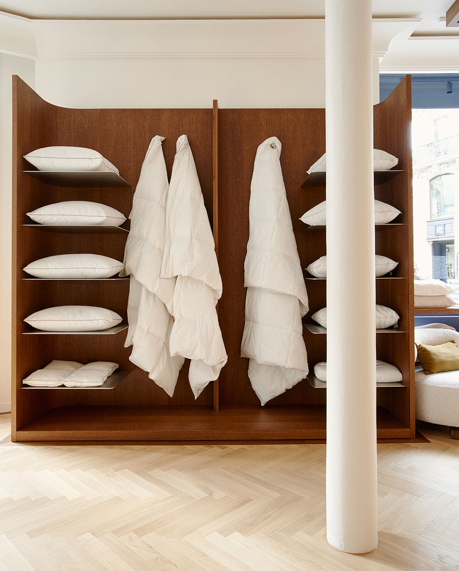 Des étagères en bois sur lesquelles sont disposés des coussins blancs et des couettes suspendues, avec une colonne blanche à l'avant, dans une pièce dotée d'un parquet en bois clair.