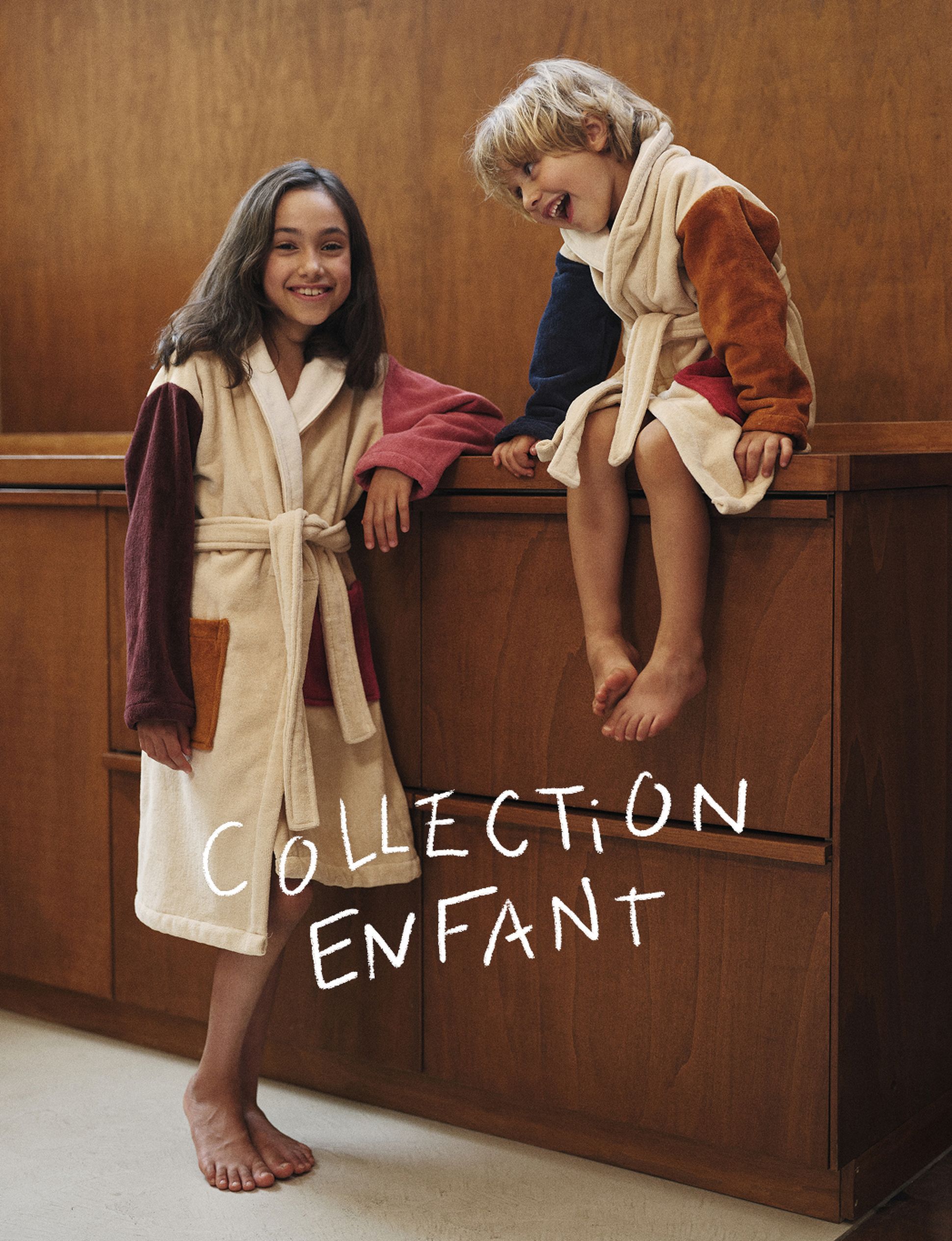 Deux enfants en peignoirs colorés, l'un debout et l'autre assis sur un meuble en bois, avec les mots « Collection Enfant » inscrits au-dessus d'eux.