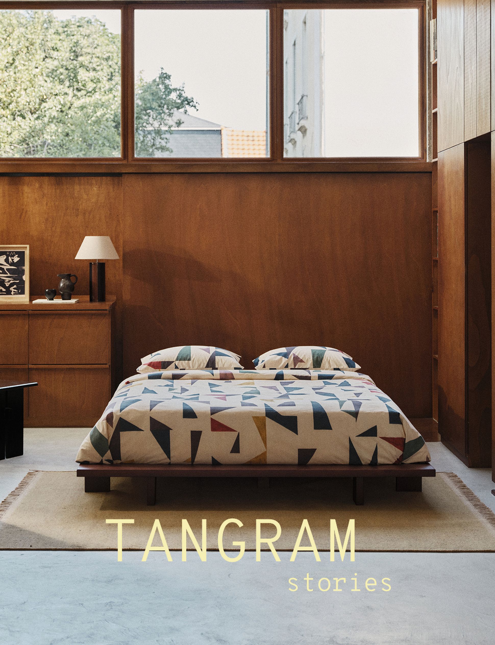 Une chambre minimaliste avec un couvre-lit à motifs géométriques, des murs en bois, de grandes fenêtres et une lampe de chevet. L'inscription « TANGRAM stories » figure sur l'image.