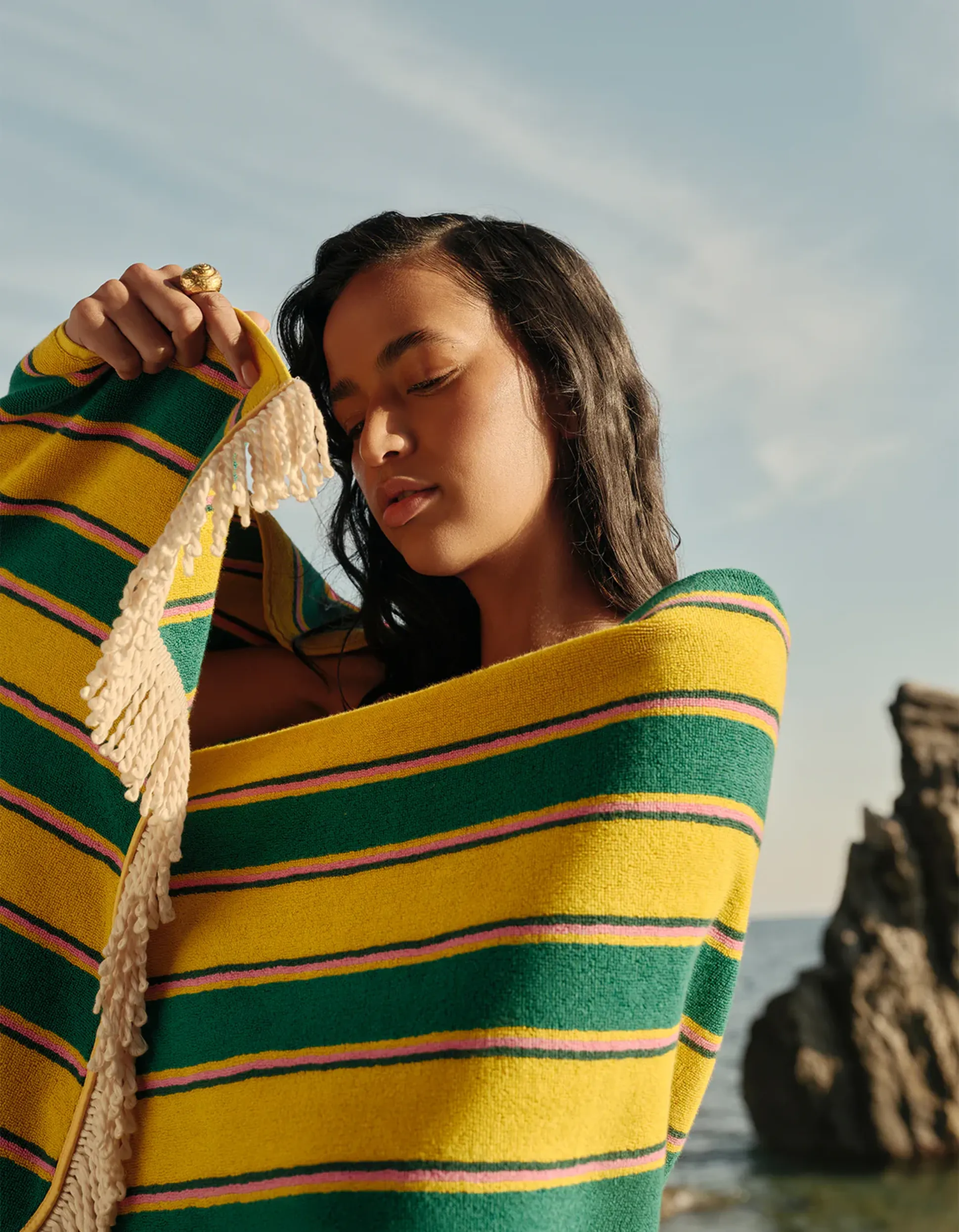 Une femme enveloppée dans une couverture rayée jaune et verte se tient debout sur une plage, les yeux fermés, avec une falaise rocheuse et l'océan en arrière-plan.