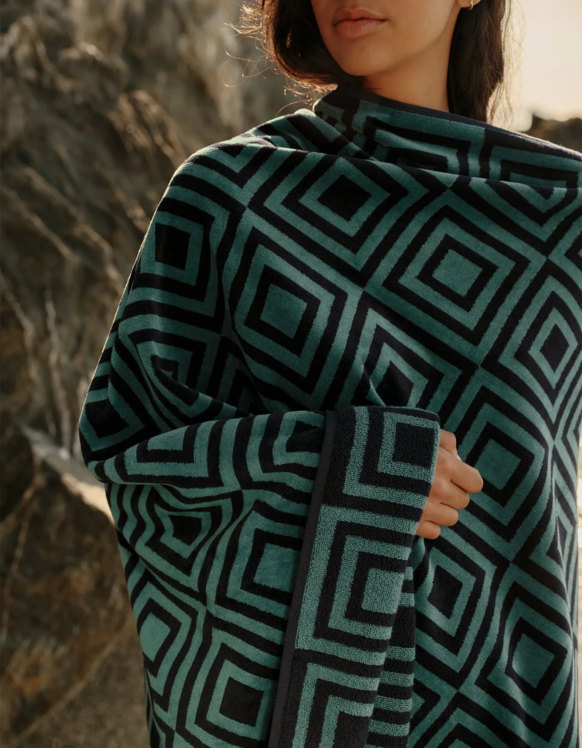 Une personne enveloppée dans une couverture à motifs géométriques, debout à l'extérieur près d'un terrain rocheux, partiellement dissimulée par les ombres.