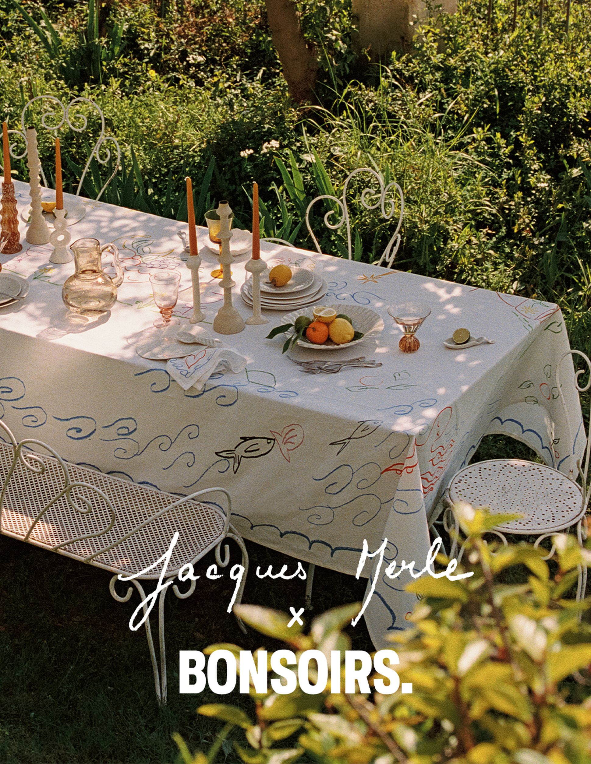 Une table d'extérieur recouverte d'une nappe à motifs, avec des bougies, des fruits et des verres, entourée d'une végétation luxuriante. Texte : « Jacques Merle x Bonsoirs. »