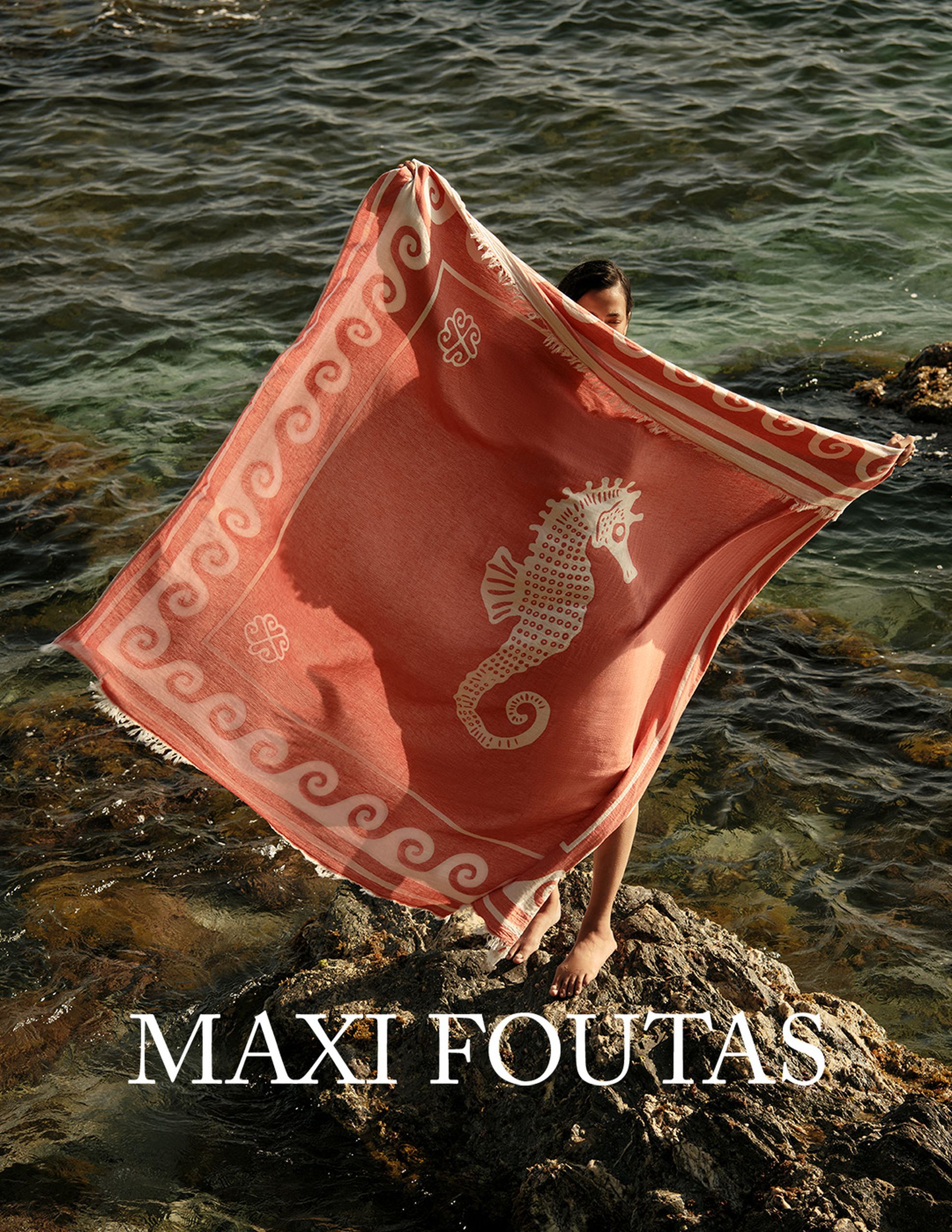 Une personne tenant une serviette de plage en corail ornée d'un motif d'hippocampe blanc, debout sur des rochers au bord de la mer. Le texte indique « MAXI FOUTAS ».