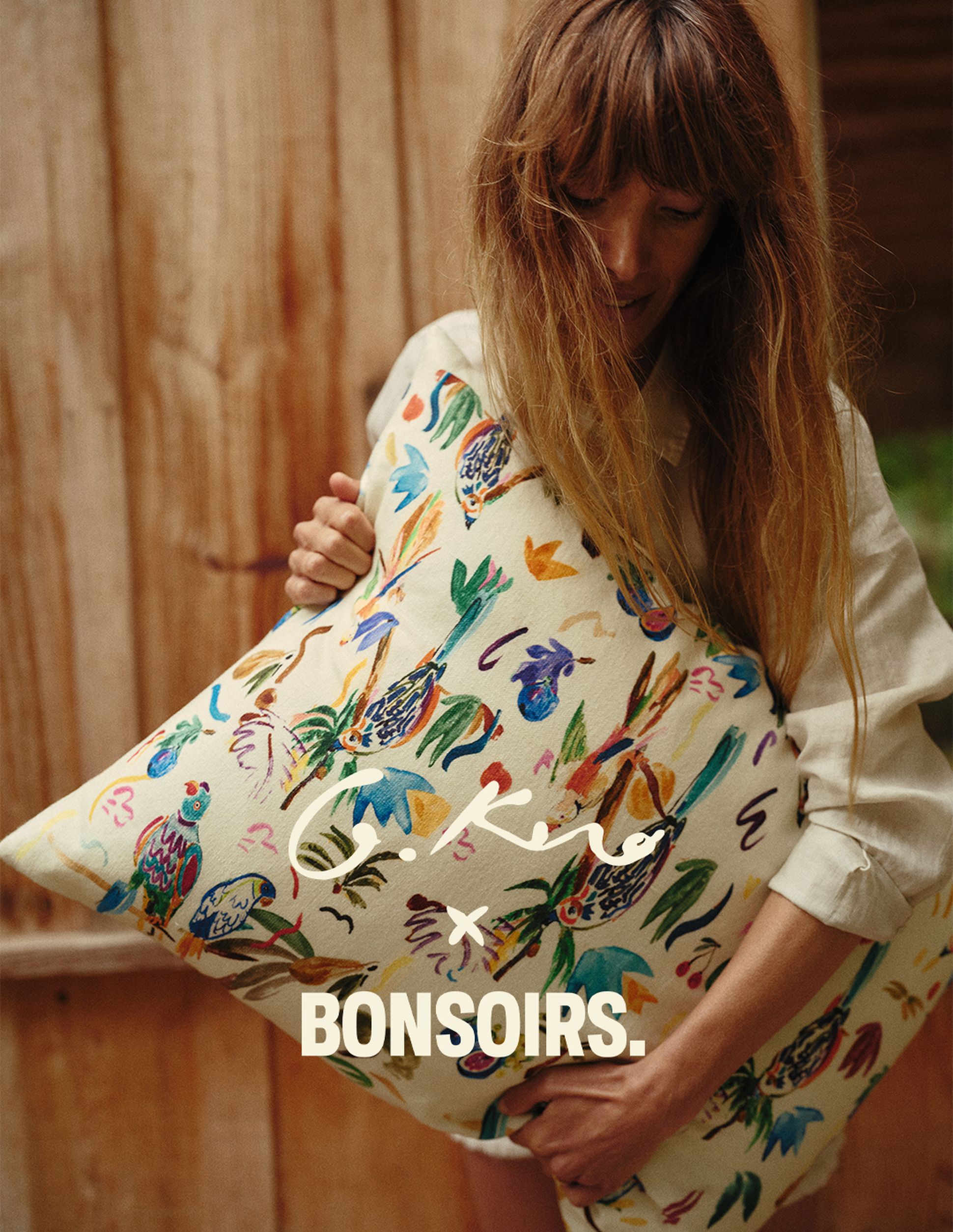 Une personne tenant un coussin coloré à motifs sur lequel figure l'inscription « G.Kero x BONSOIRS », devant un fond en bois.