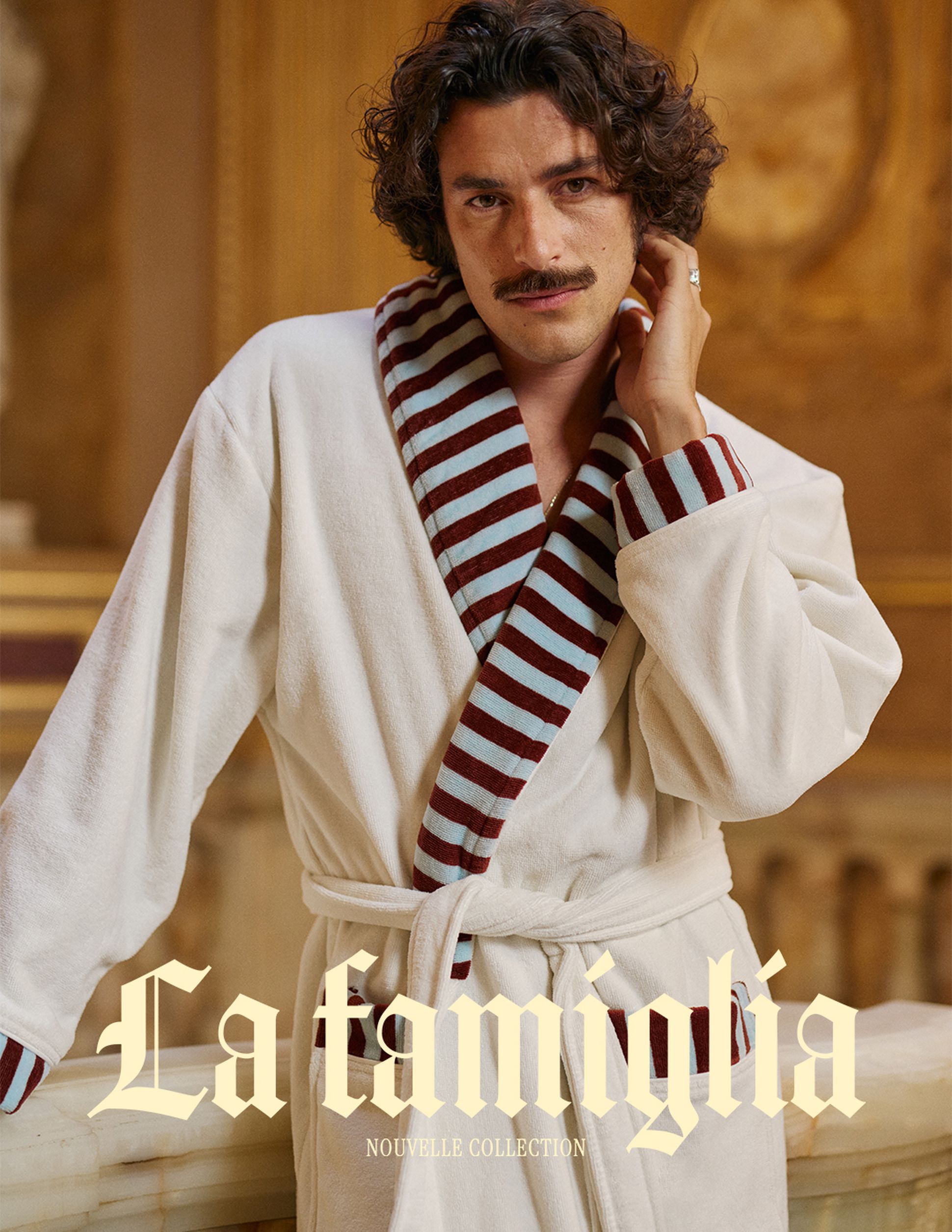 Un homme vêtu d'un peignoir au col rayé pose avec assurance, la main posée sur son cou. Le texte indique « La famiglia : Nouvelle collection ». Un décor élégant sert de toile de fond.