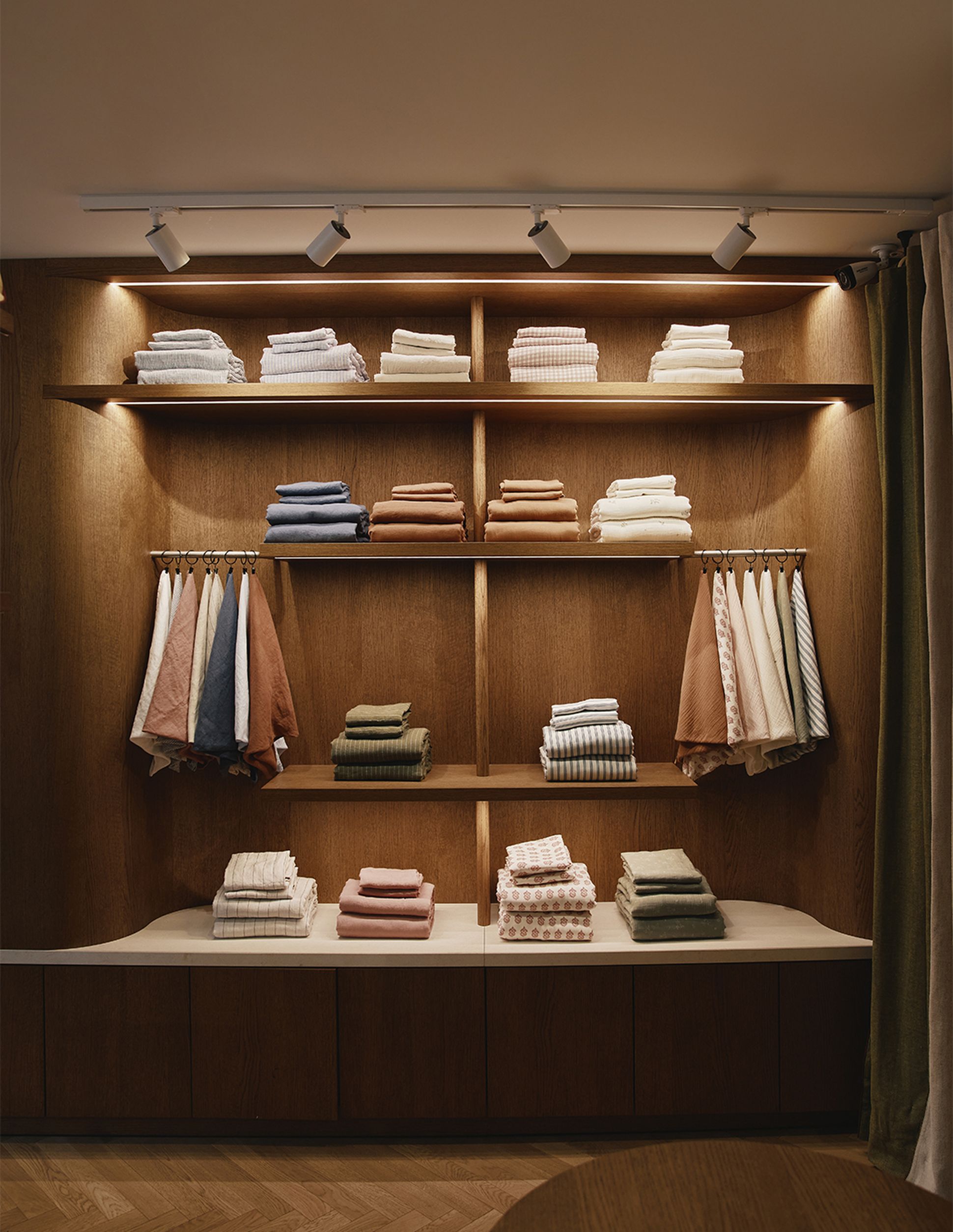 Une étagère en bois sur laquelle sont soigneusement empilées des serviettes et suspendus des peignoirs, le tout éclairé par des spots, dans un cadre moderne et chaleureux.