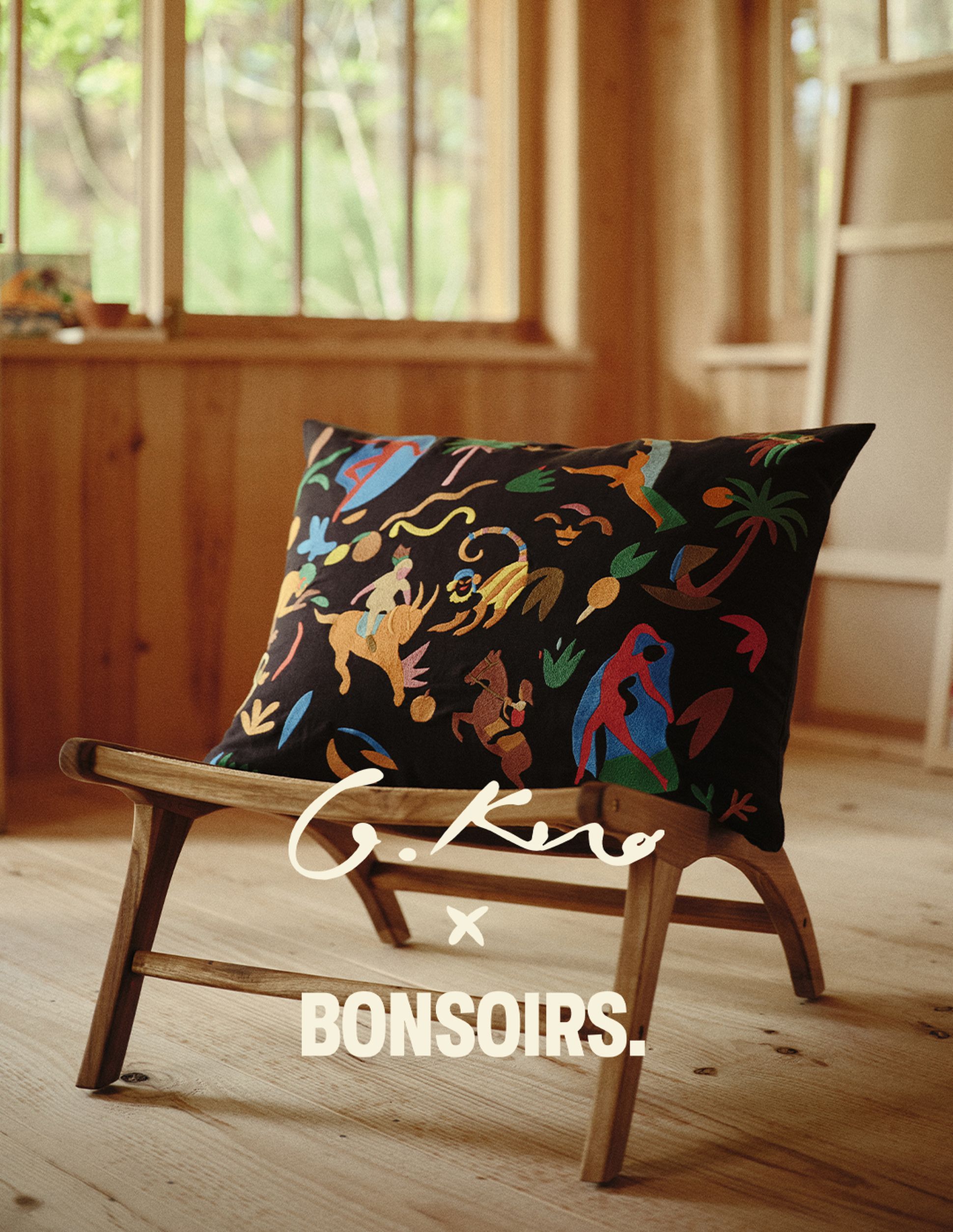 Un coussin décoratif orné de motifs d'animaux colorés, posé sur une chaise en bois dans une pièce chaleureuse dotée de grandes fenêtres. On peut y lire : « G. Kero x Bonsoirs ».