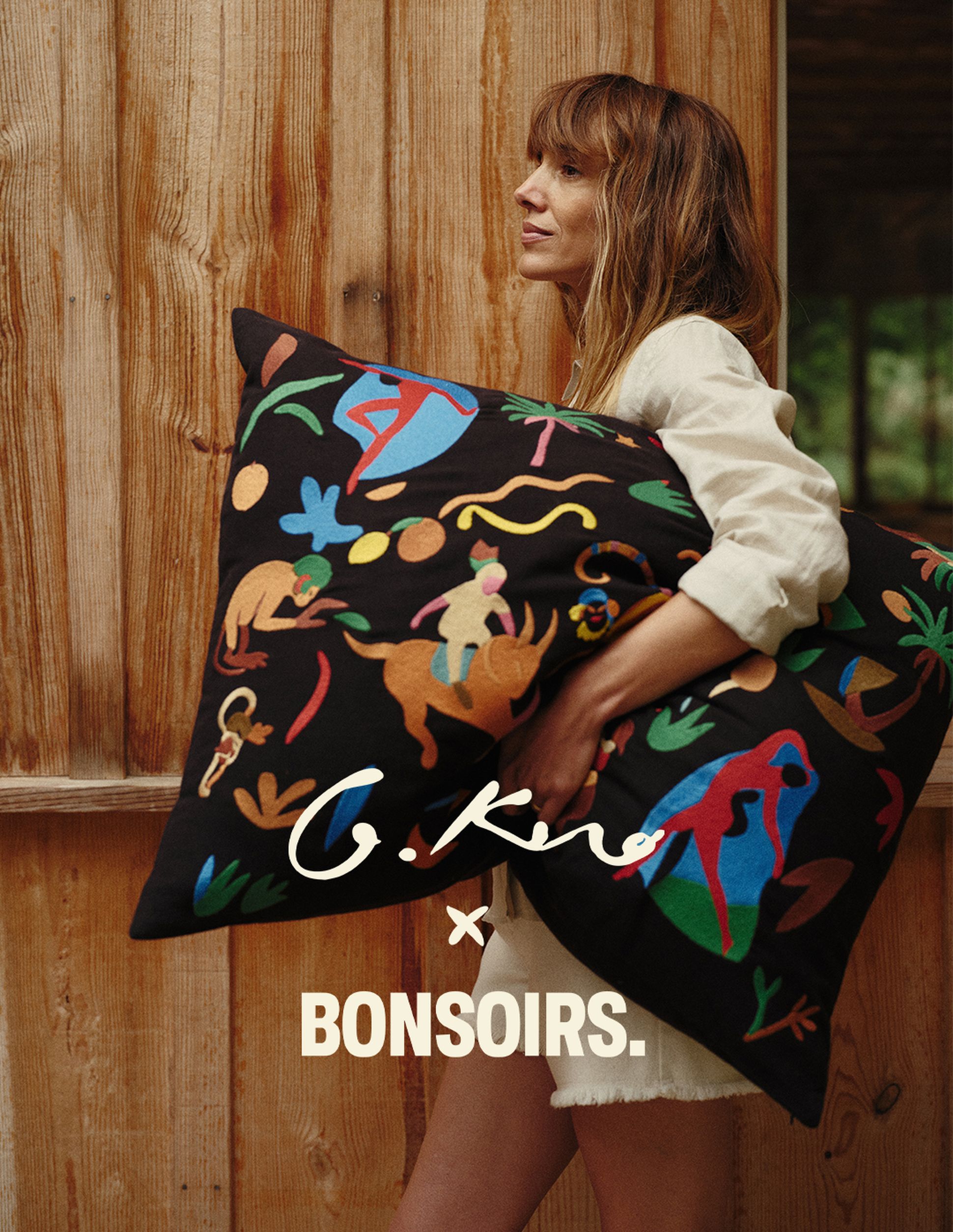 Une femme tenant un coussin coloré à motifs devant un mur en bois, sur lequel figure l'inscription « G.Kero x Bonsoirs ».