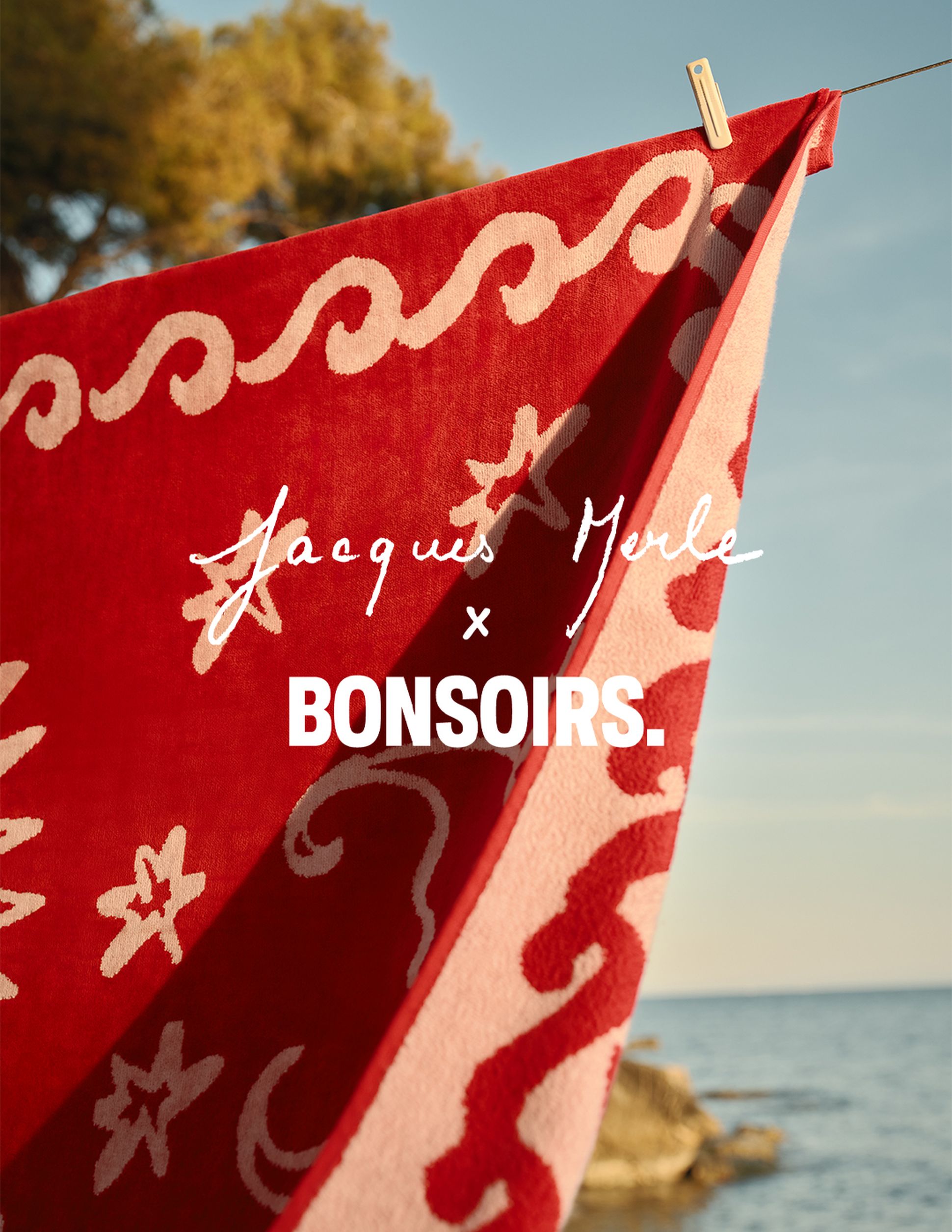 Une serviette rouge ornée de motifs ondulés blancs, suspendue à une corde au bord de la mer, sous un ciel dégagé. Légende : « Jacques Merle x BONSOIRS. »