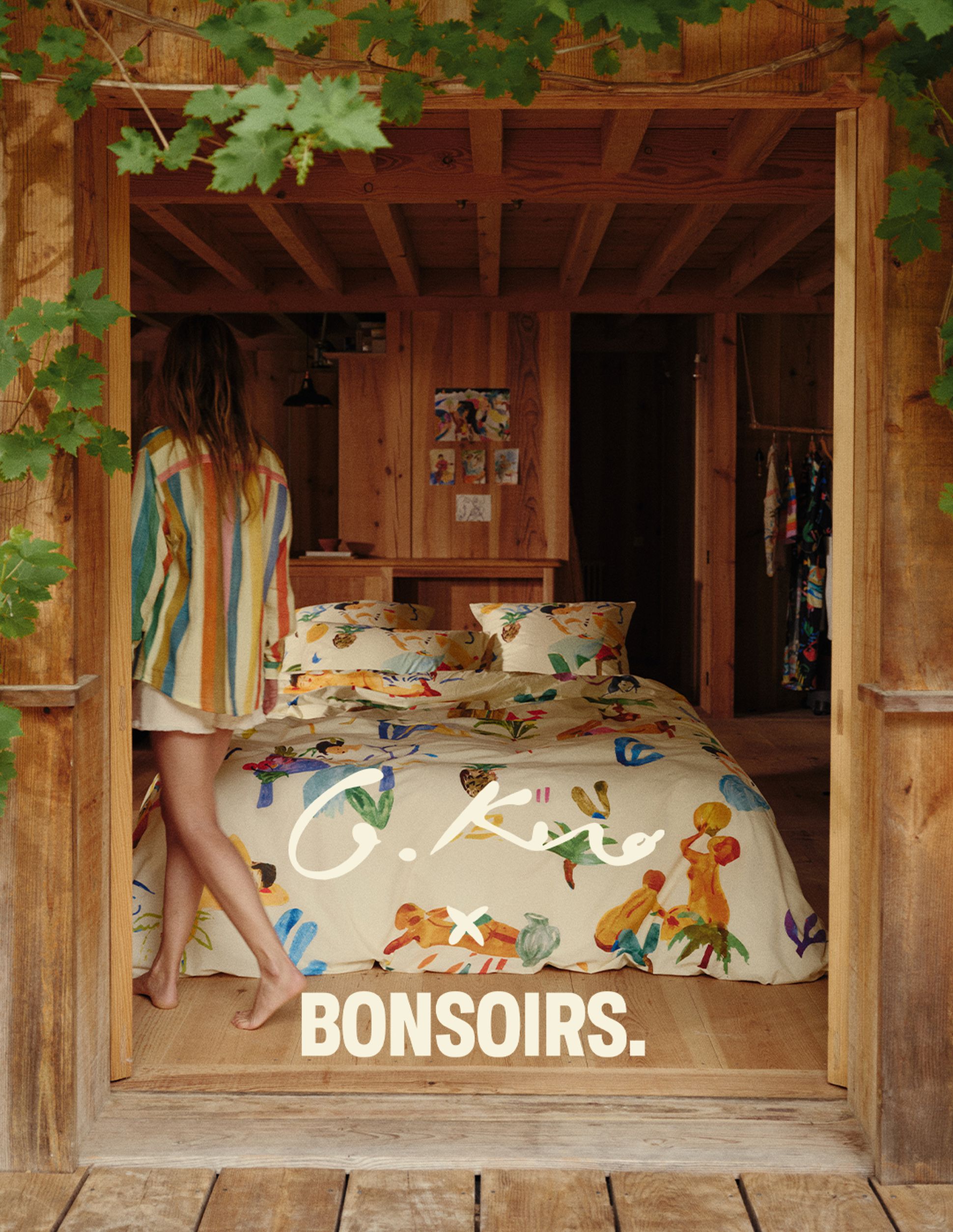 Une femme vêtue d'une chemise colorée se tient près d'un lit orné d'une parure de lit originale, dans une chambre rustique en bois, sur laquelle est inscrit « G. Kero x Bonsoirs ».