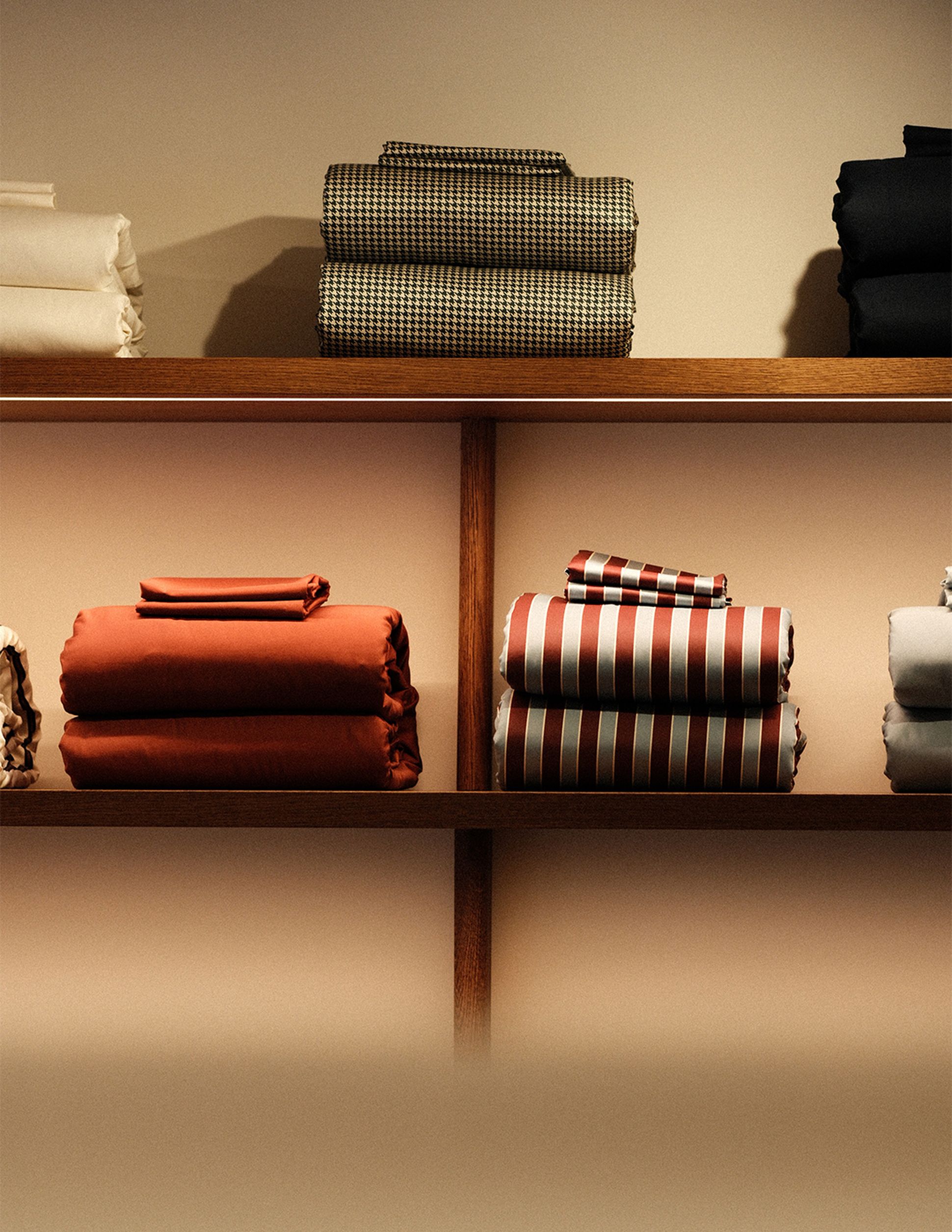 Des tissus empilés et pliés, de différentes couleurs et motifs, présentés sur des étagères en bois, notamment des modèles à rayures et à carreaux.