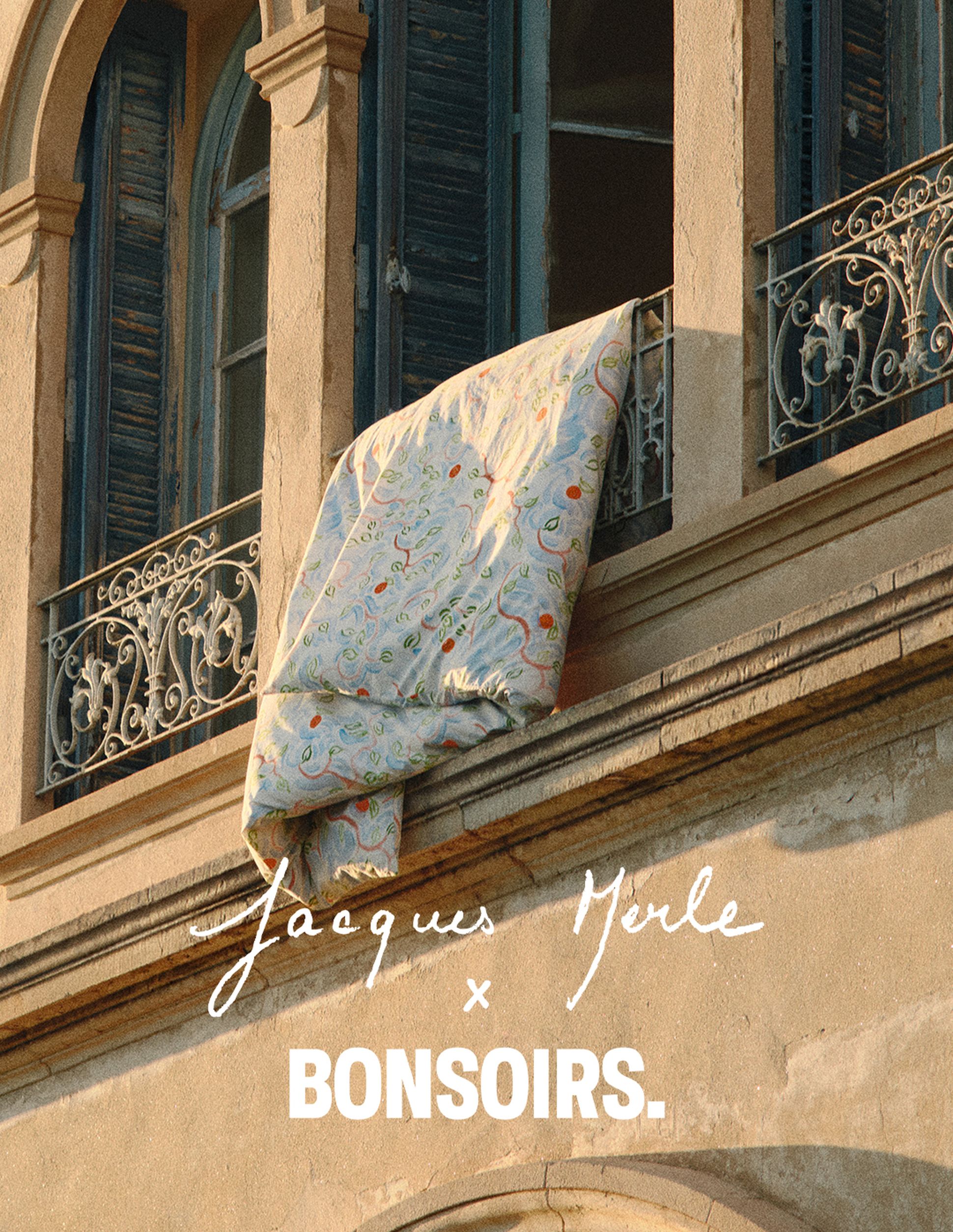 Une courtepointe est suspendue au balcon décoratif d'un immeuble en pierre aux volets bleus, sur laquelle on peut lire : « Jacques Merle x Bonsoirs ».
