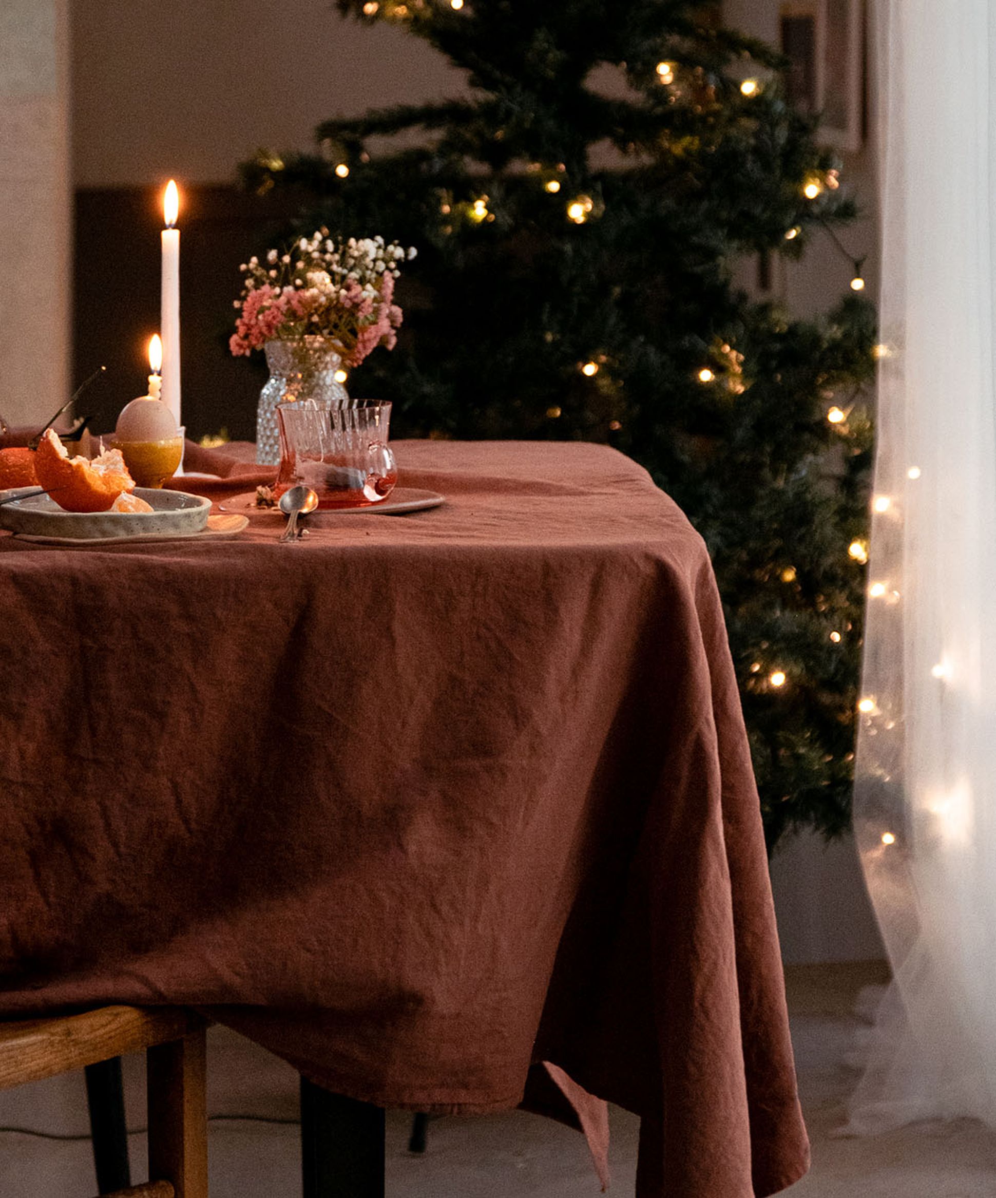 Ambiance de table chaleureuse avec une nappe marron, une bougie allumée, des fleurs, de la verrerie, et un sapin de Noël décoré de lumières chaudes en arrière-plan.
