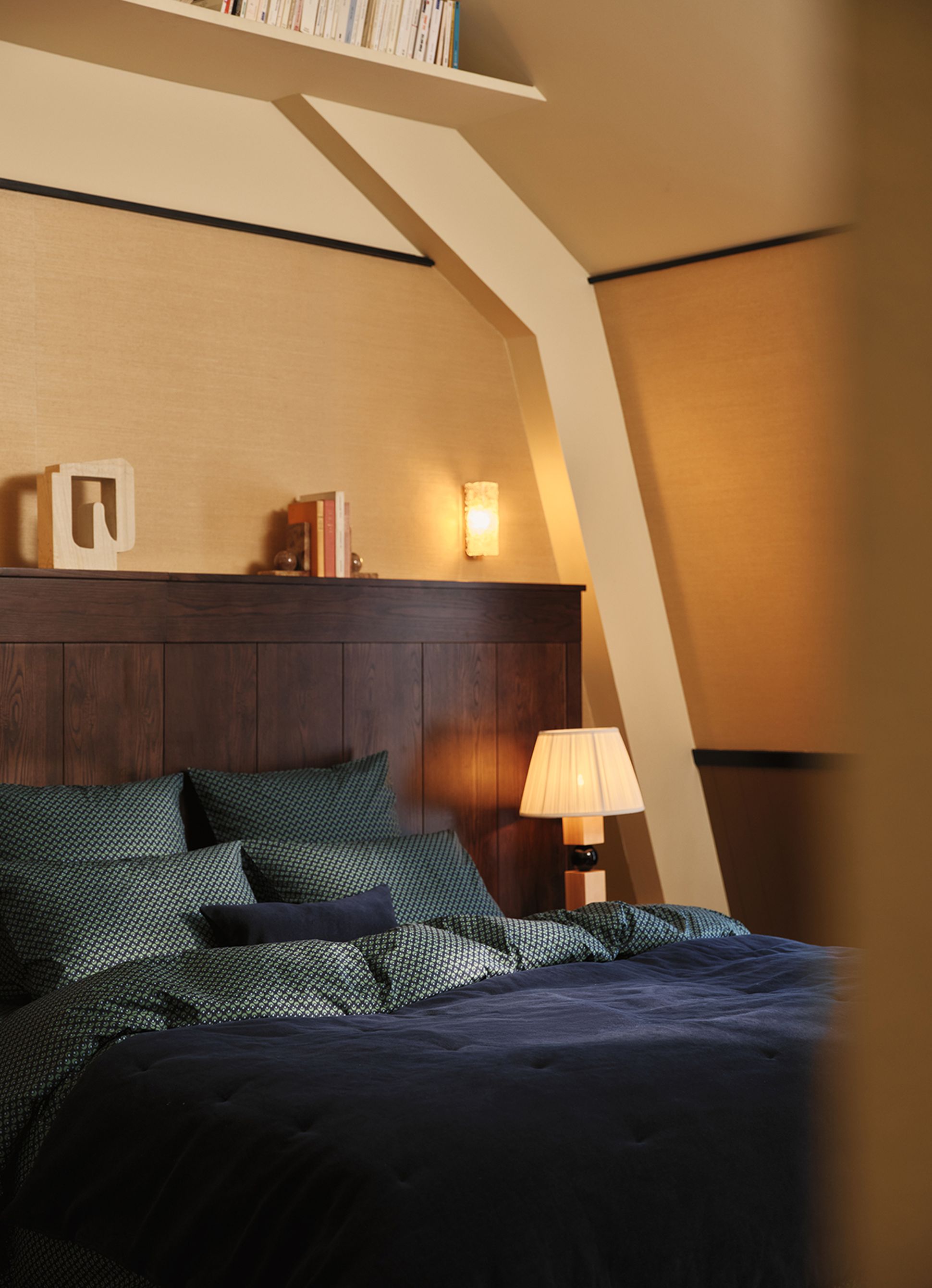 Cozy bedroom with a wooden headboard, patterned pillows, a navy duvet, and warm lighting. A shelf with books decorates the angled ceiling.