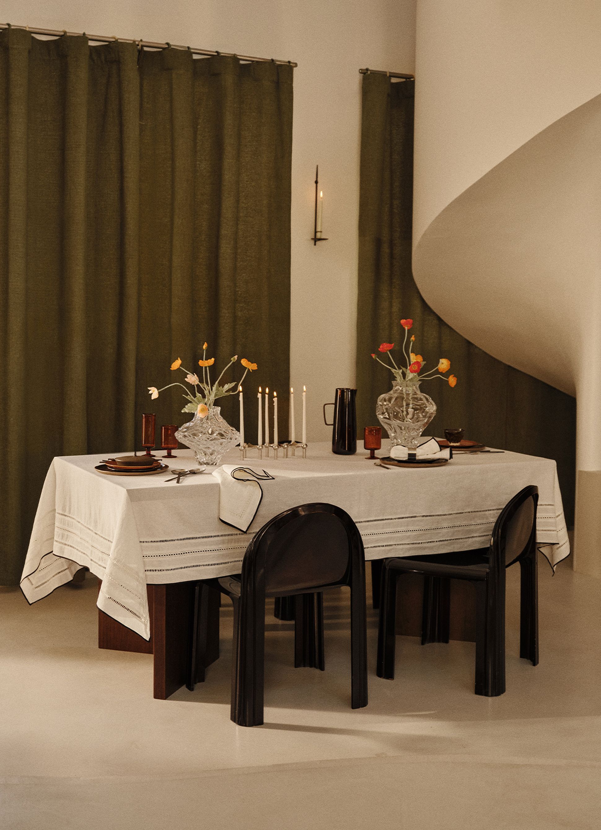 Une élégante table à manger dressée avec une nappe en lin blanc, des vases en cristal remplis de fleurs, des bougeoirs et des chaises foncées, le tout sur fond de rideaux vert olive.