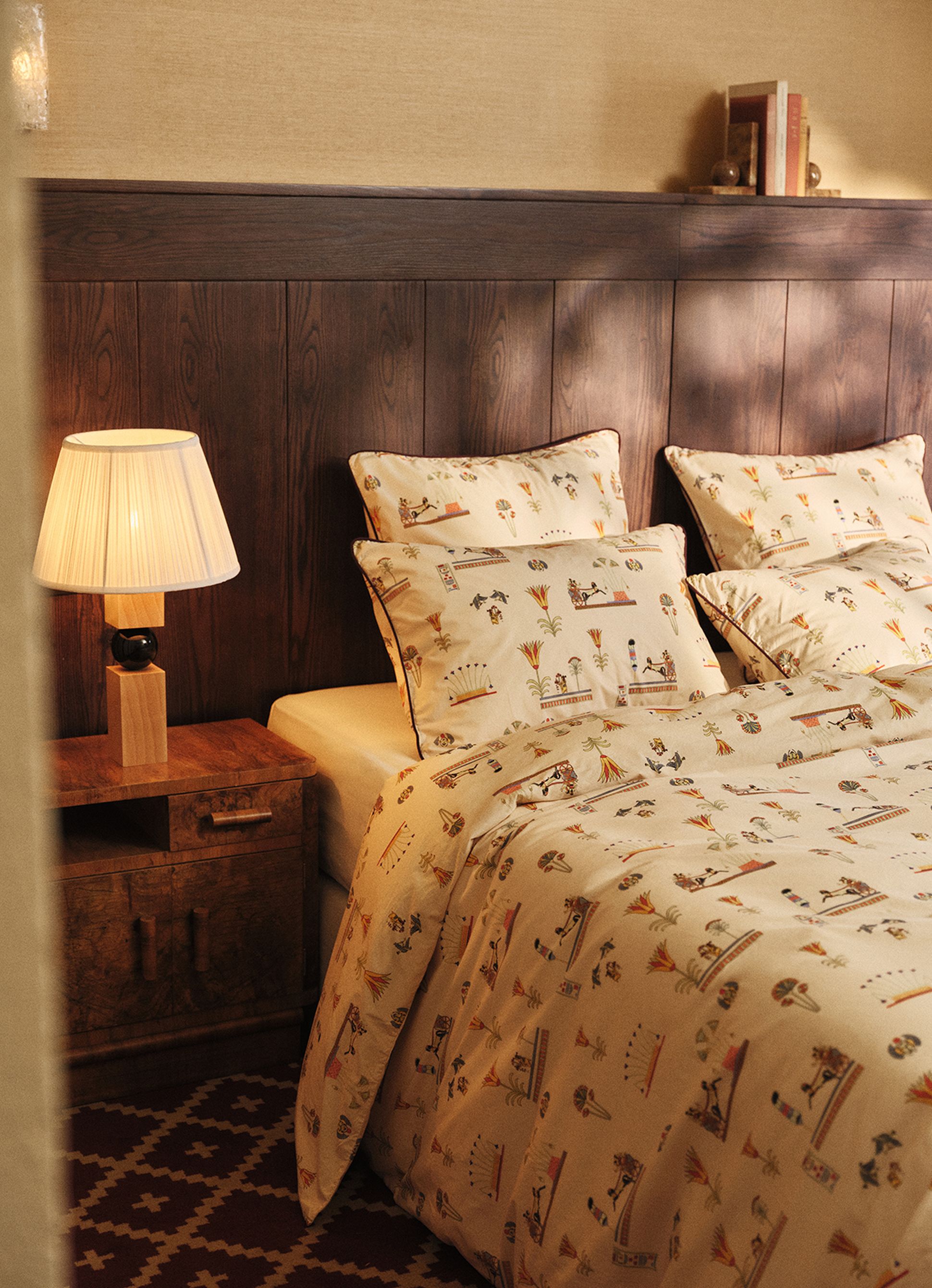 Cozy bedroom with a wooden headboard, patterned bedding featuring Egyptian motifs, a wooden side table, and a lamp with a pleated shade.