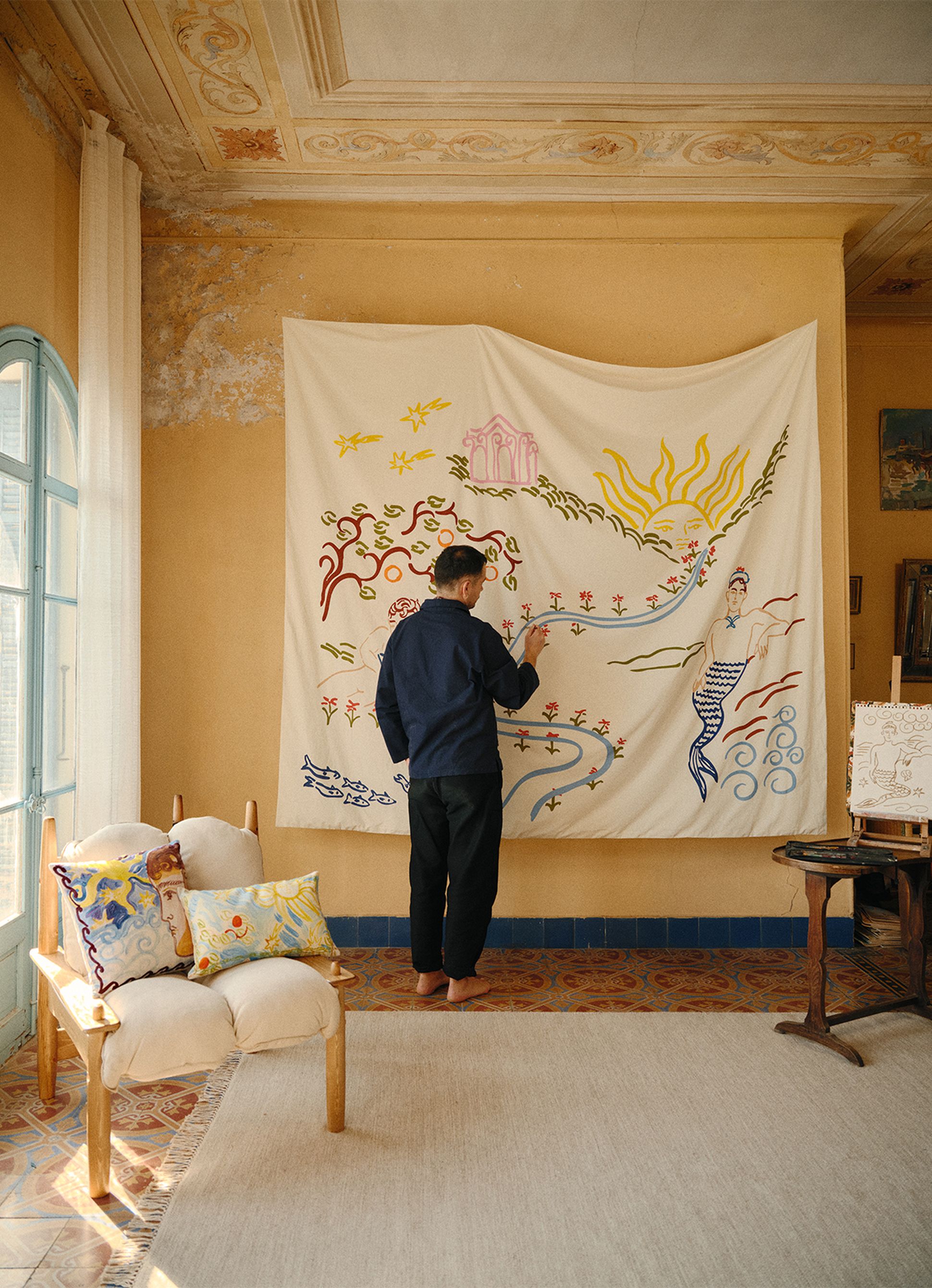 Une personne en train de peindre une grande fresque murale colorée sur un mur, dans une pièce chaleureuse dotée d'un sol à motifs, d'une chaise ornée de coussins décoratifs et d'un chevalet.