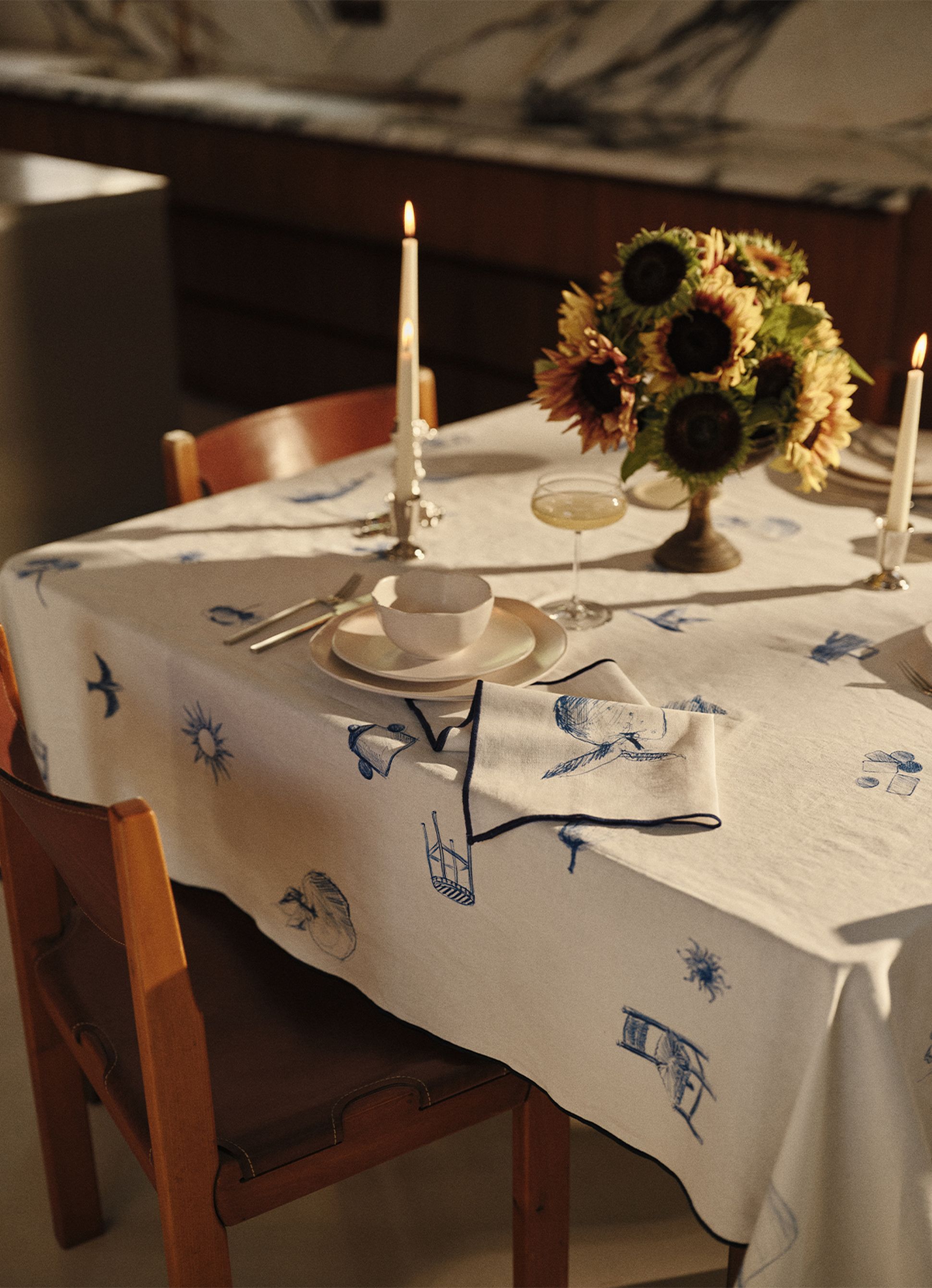 Une table éclairée à la lueur des bougies, recouverte d'une nappe blanche à motifs bleus, dressée avec des assiettes, une tasse, des couverts, une coupe à champagne et un bouquet de tournesols.