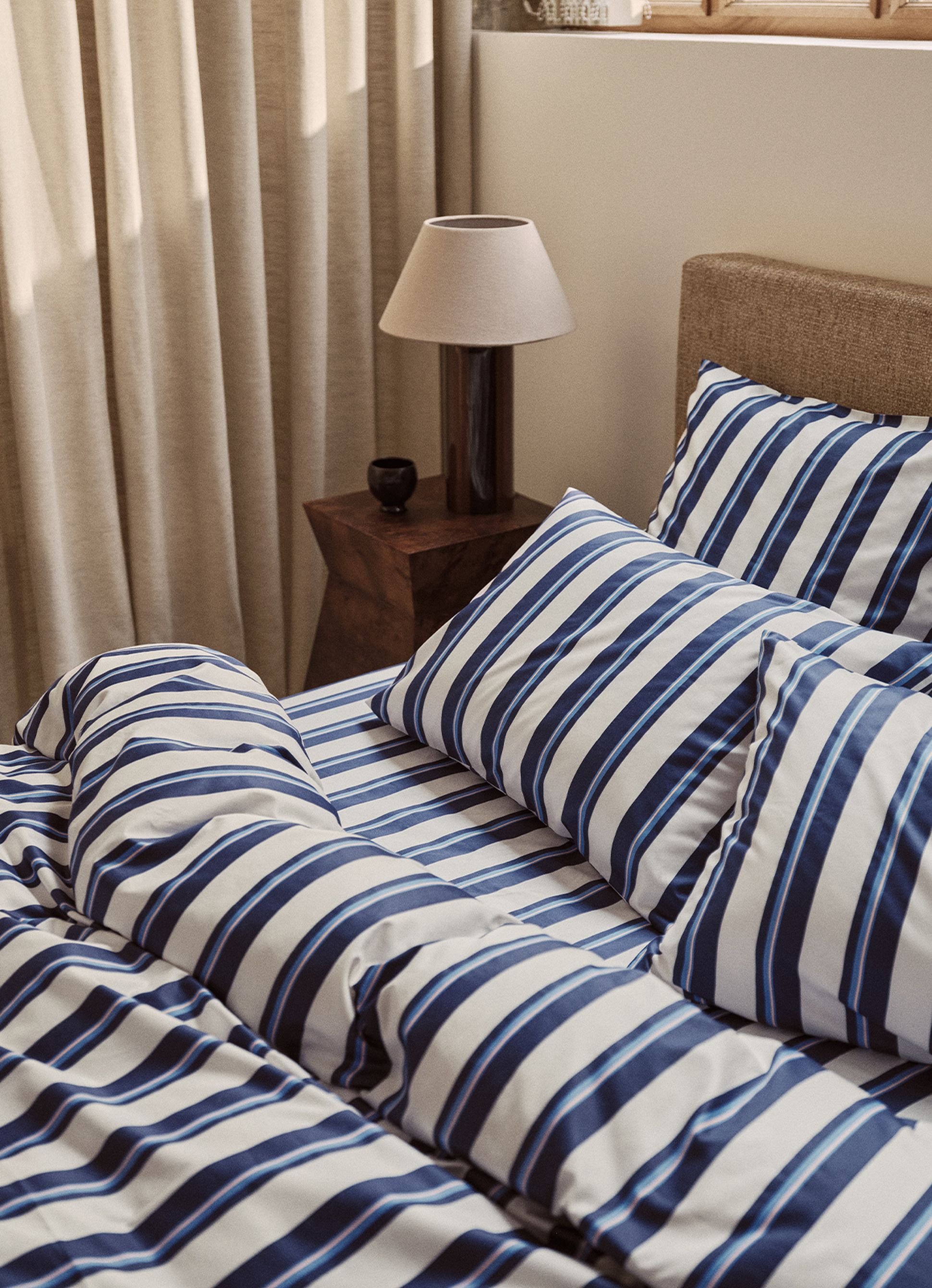 Un lit soigneusement fait, avec une parure de lit à rayures bleues et blanches, à côté d'une table de chevet en bois sur laquelle se trouvent une lampe et un petit bol, avec des rideaux en arrière-plan.
