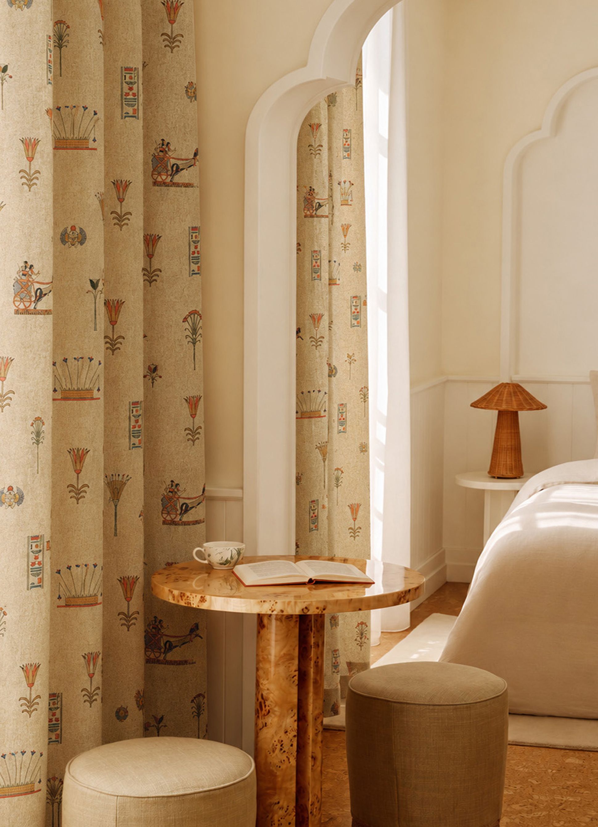 Cozy room with patterned curtains, an arched window, a small round table with a book and teacup, and a bed beside a retro lamp on a bedside table.