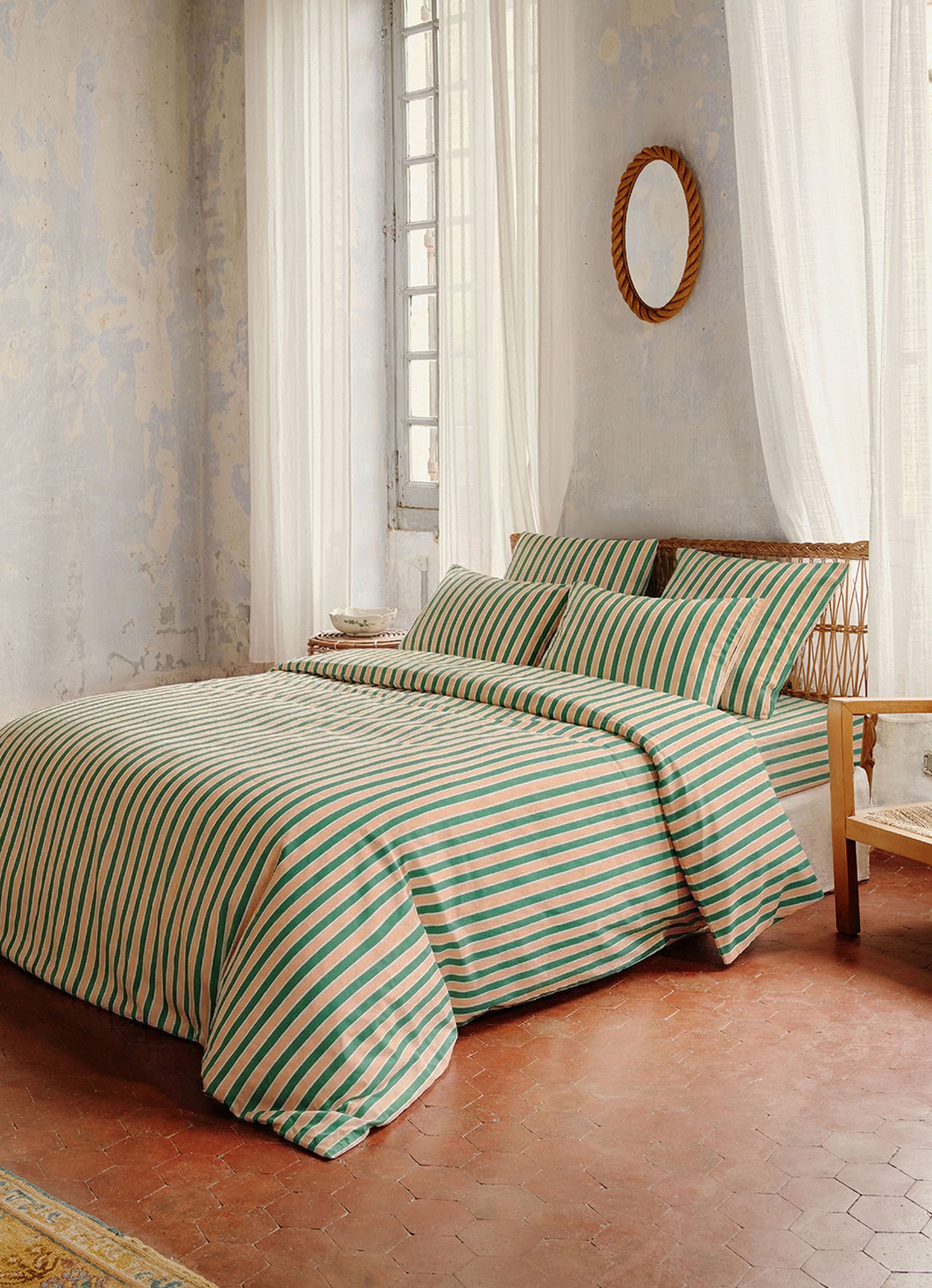 Une chambre douillette avec un ensemble de literie à rayures vertes, un lit en osier et des rideaux voilés. Un miroir rond accroché à un mur rustique met en valeur ce décor plein de charme.
