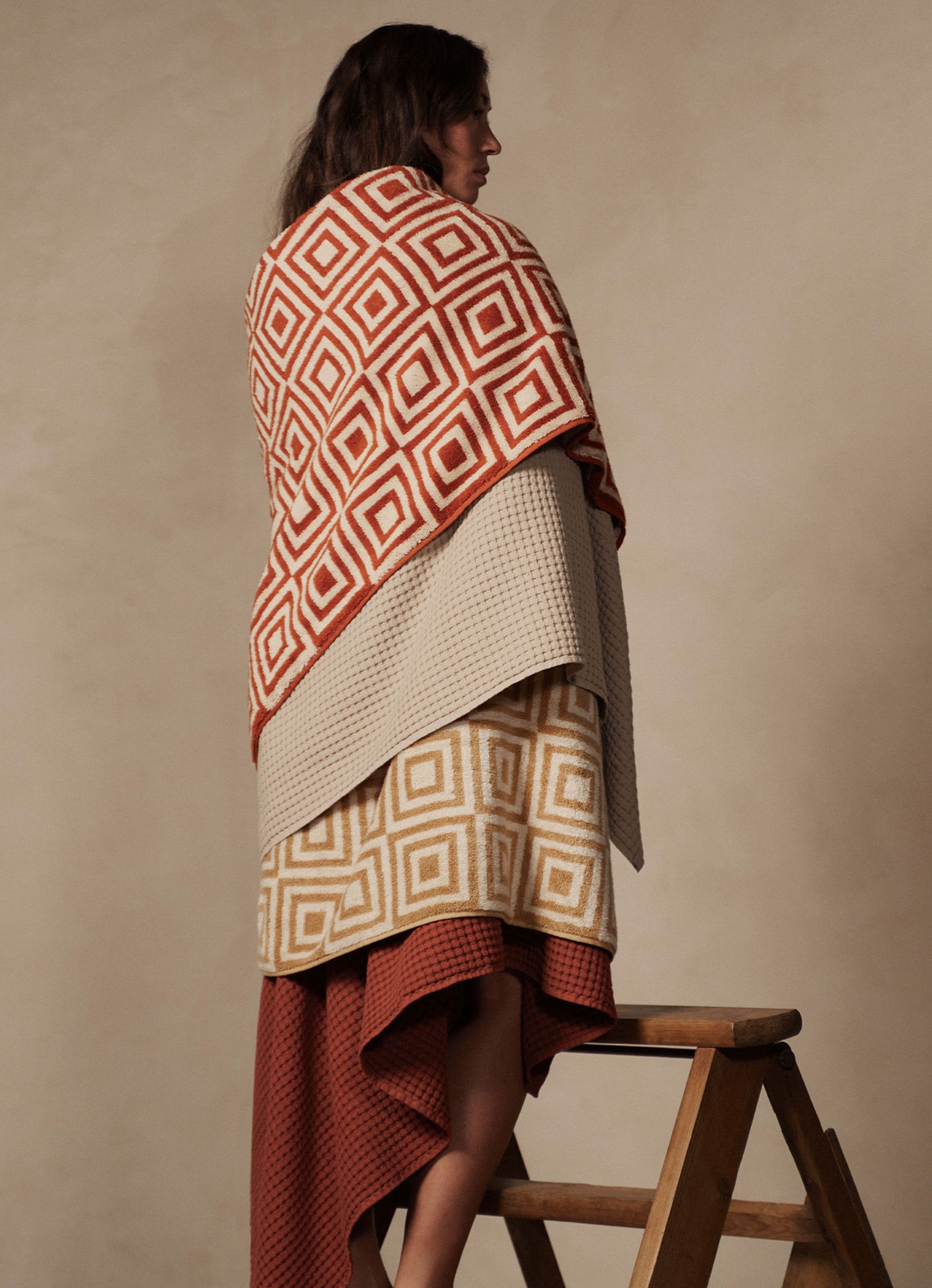 Une personne enveloppée dans des couvertures à motifs géométriques se tient debout sur une échelle en bois devant un fond neutre.