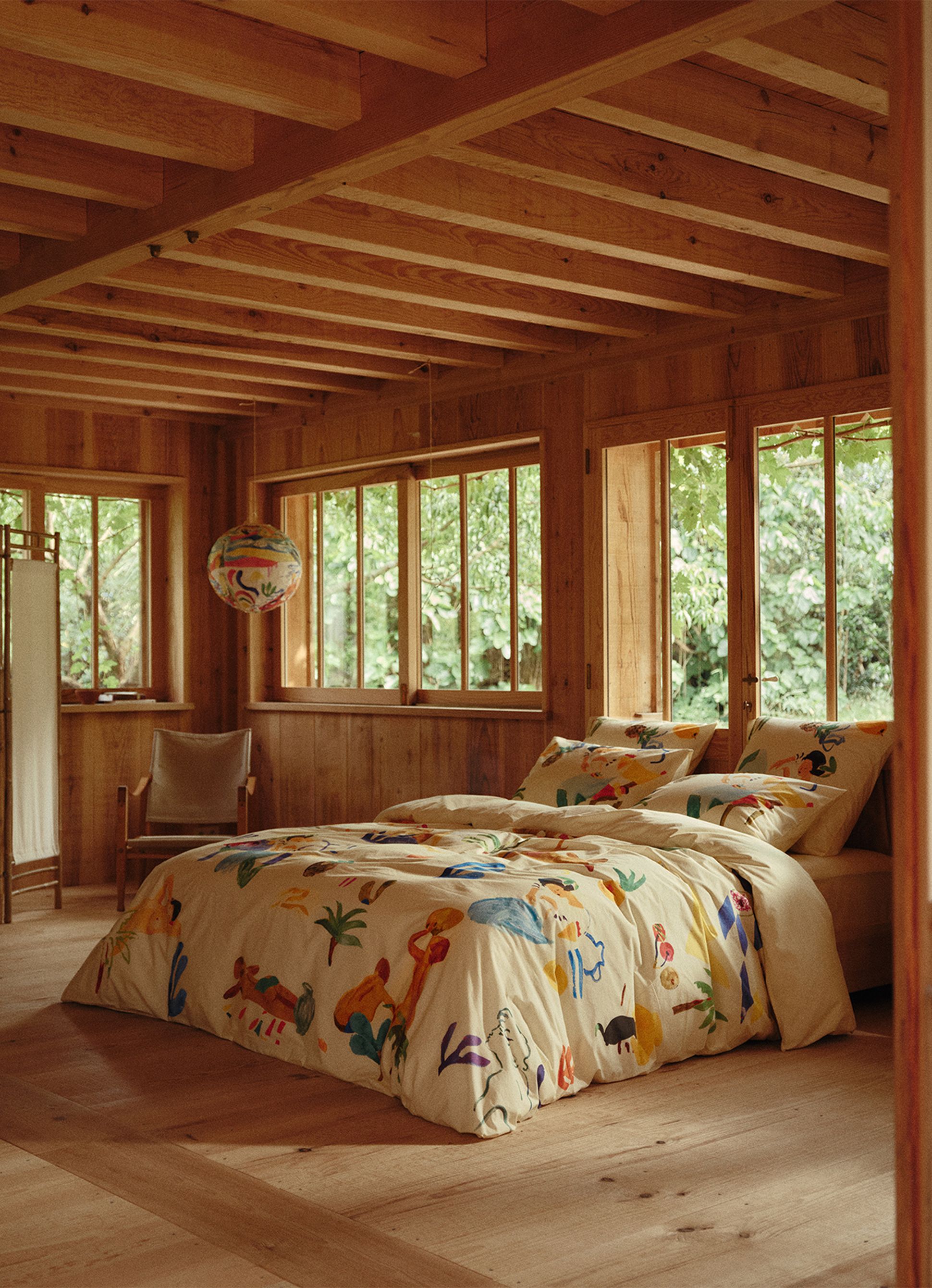 Linge de lit décoratif orné d'illustrations colorées représentant des personnages, des fruits et des plantes, le tout dans une chambre chaleureuse en bois dotée d'une fenêtre et d'un tabouret.