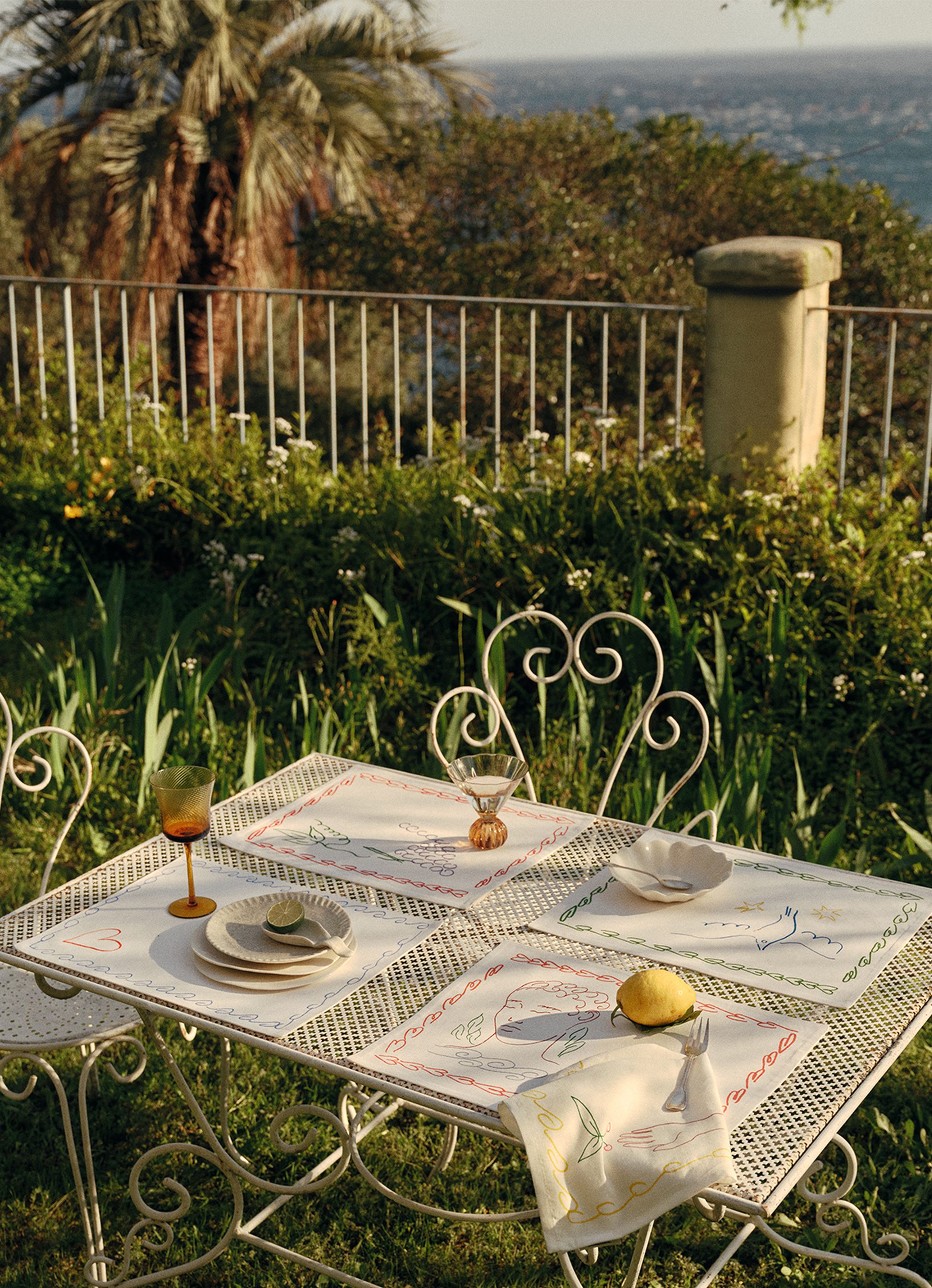 Une table de jardin dressée pour deux, avec des sets de table décorés, des verres et un citron, offrant une vue panoramique sur des palmiers et de la verdure.