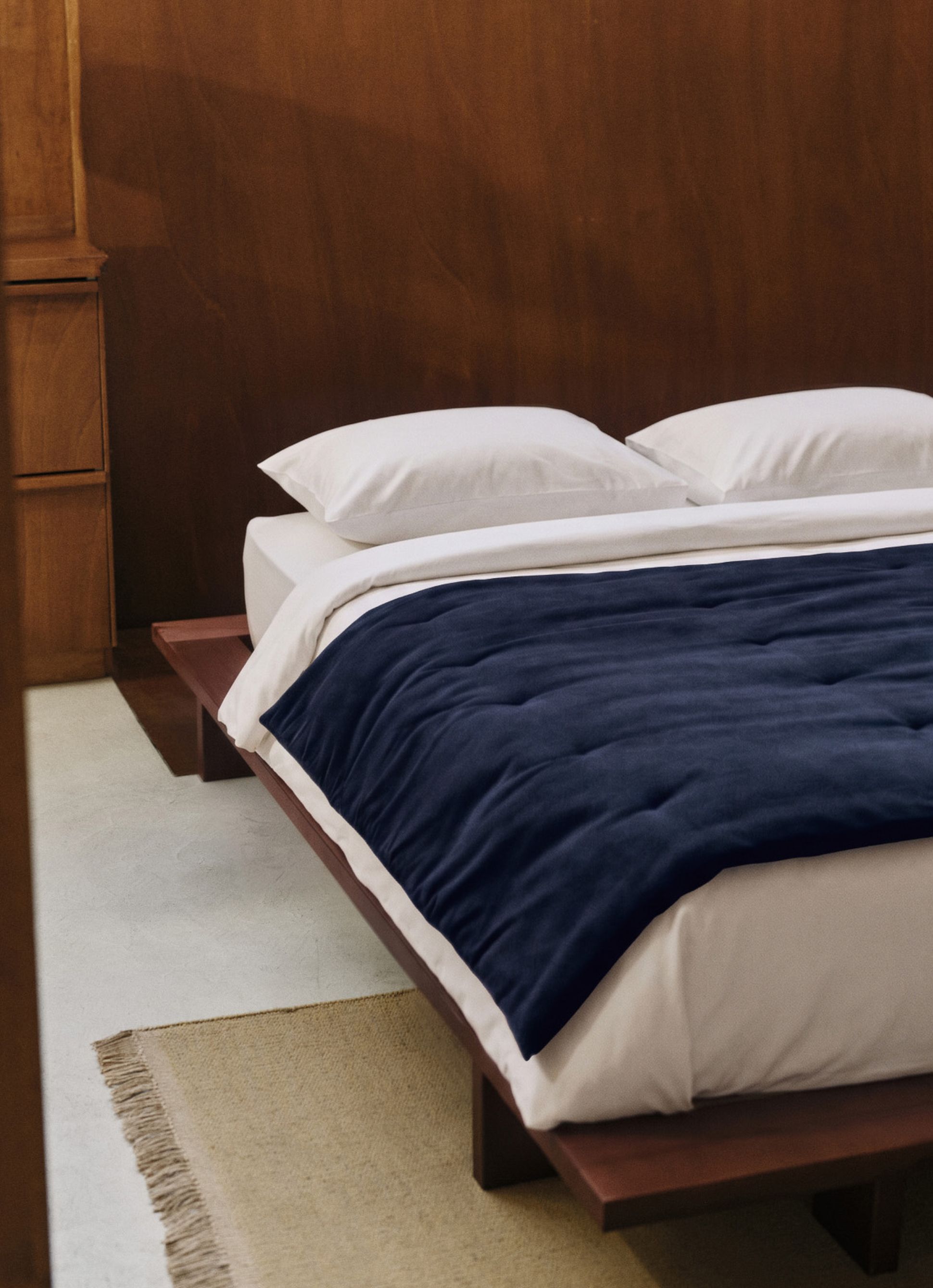 A minimalist bedroom features a wooden platform bed with crisp white bedding and a navy blue blanket, set against wood-paneled walls.