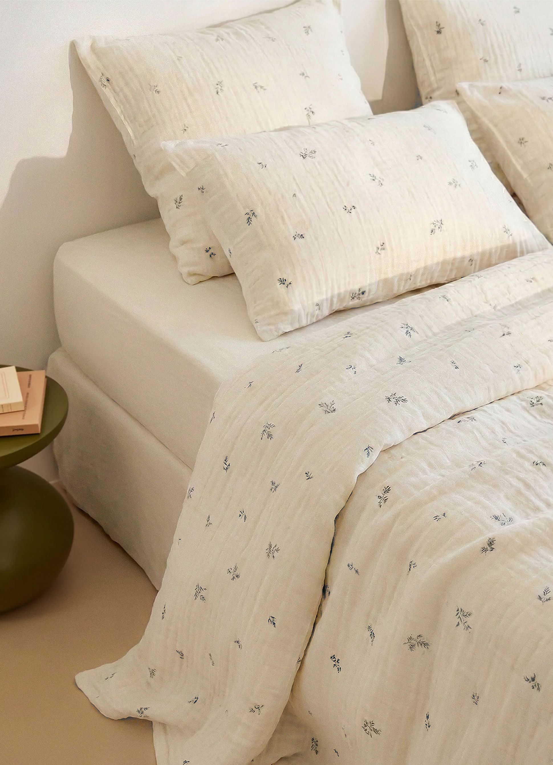 Un lit avec une literie de couleur crème ornée d'un subtil imprimé floral. L'ensemble comprend des oreillers, une couverture et des draps sur un cadre simple.