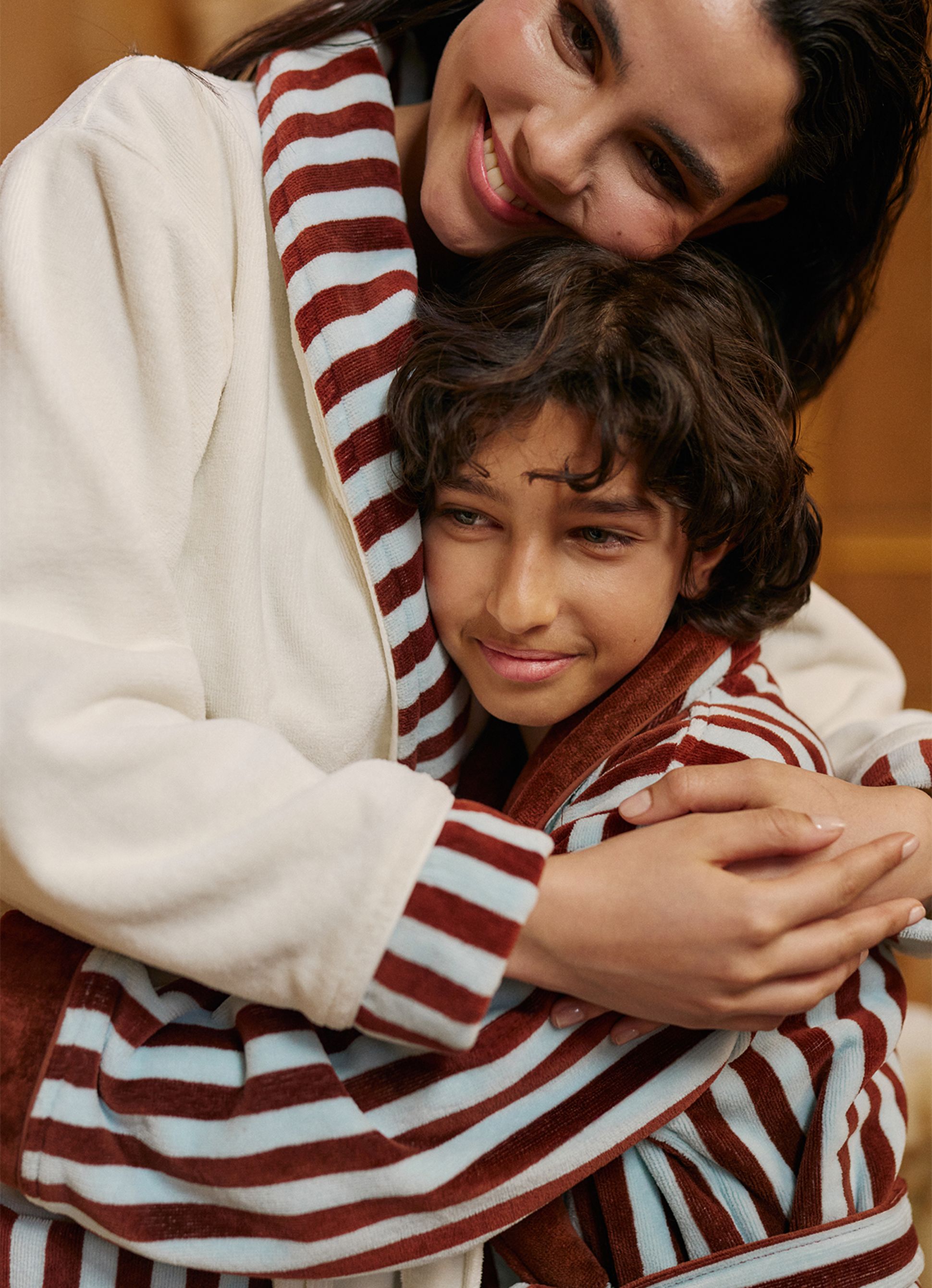 Une femme vêtue d'un peignoir rayé serre dans ses bras un enfant souriant qui porte un peignoir assorti, dans une scène empreinte de chaleur et d'affection.