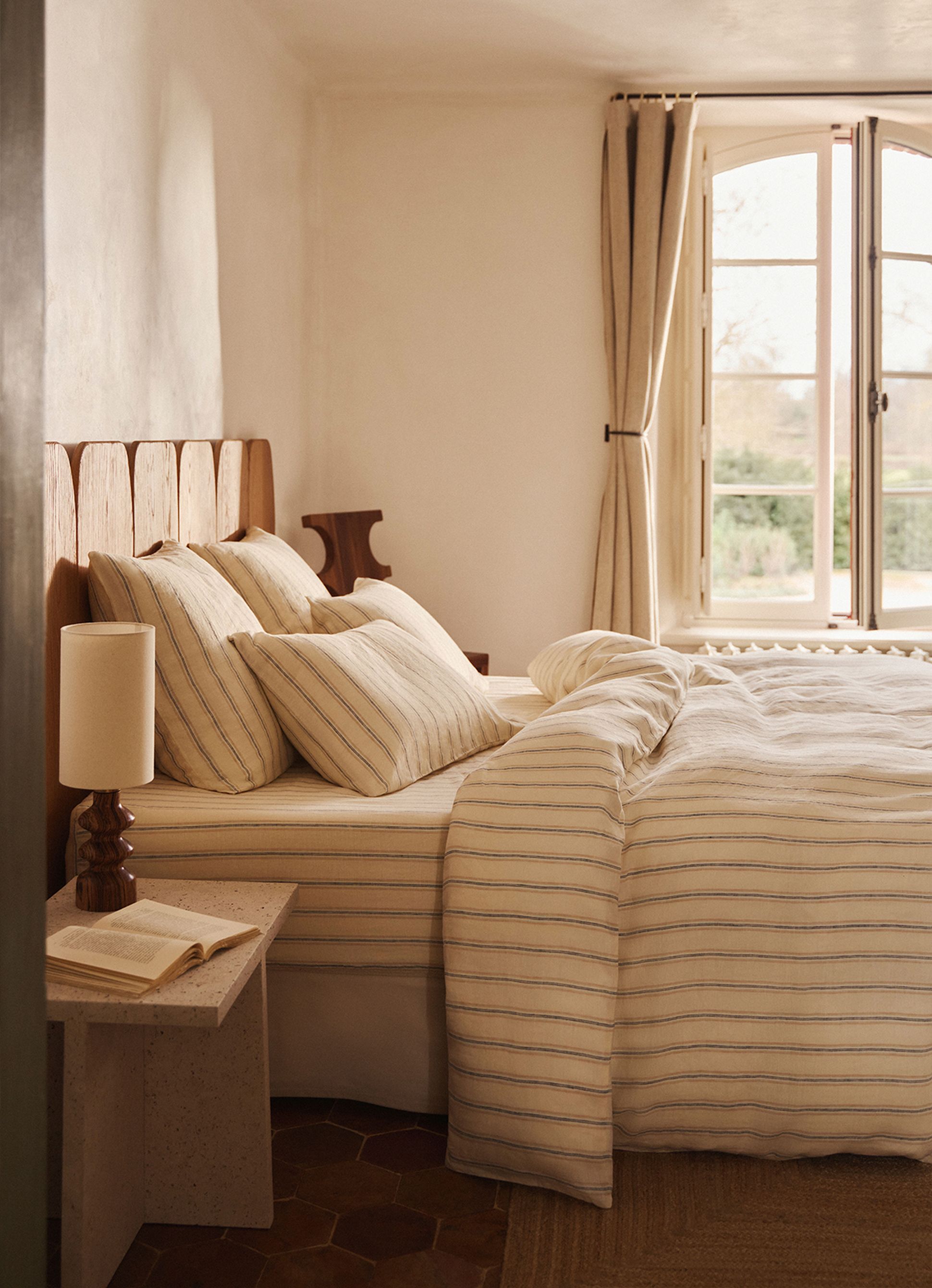 Chambre à coucher chaleureuse avec une parure de lit rayée, une tête de lit en bois, une table de chevet sur laquelle se trouvent une lampe et un livre ouvert, ainsi qu'une fenêtre aux rideaux ouverts.