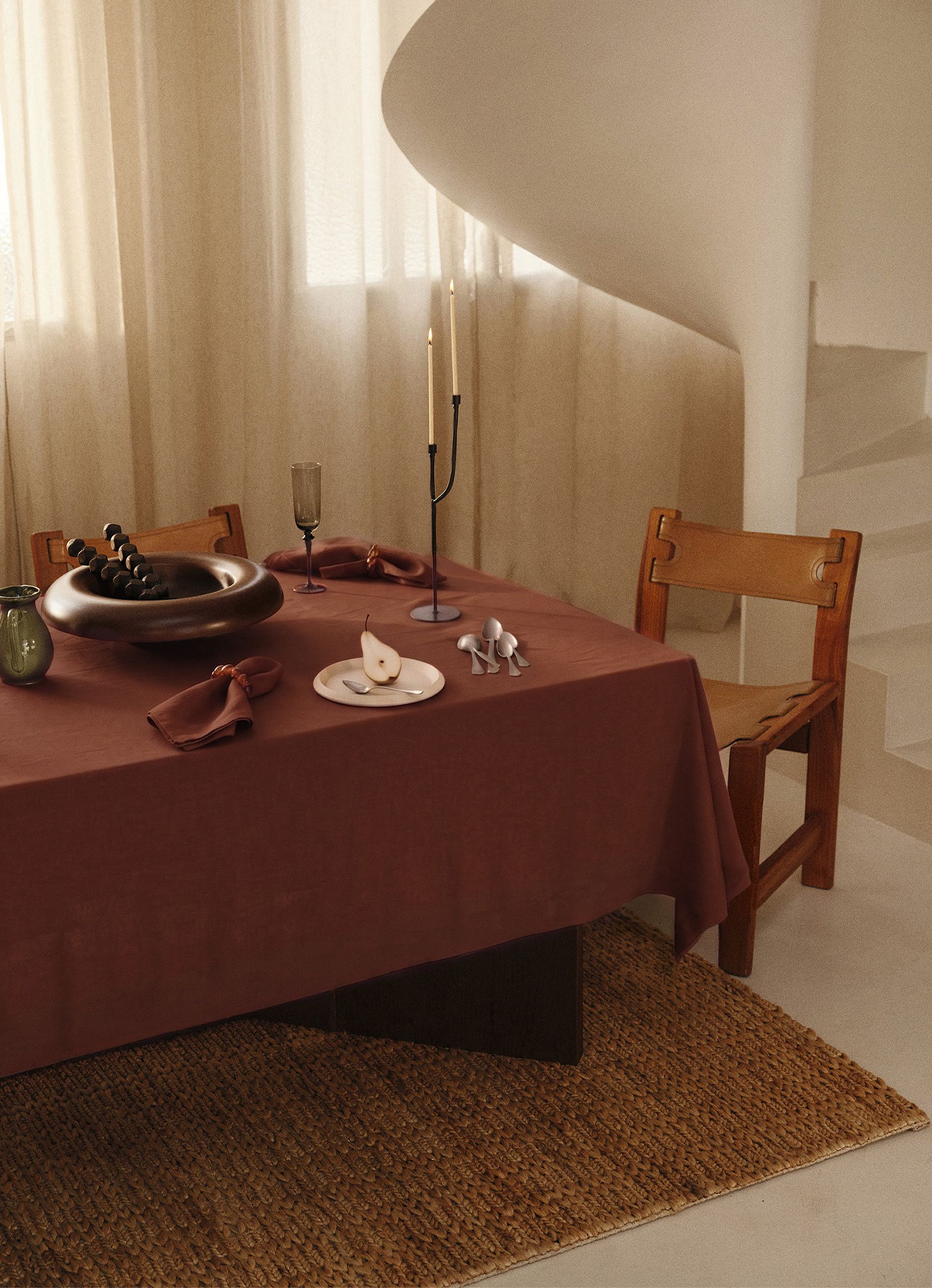 Une table à manger élégamment dressée, recouverte d'une nappe marron, avec deux grandes bougies, une poire posée sur une assiette et un bol décoratif, à côté d'une chaise en bois posée sur un tapis tissé.