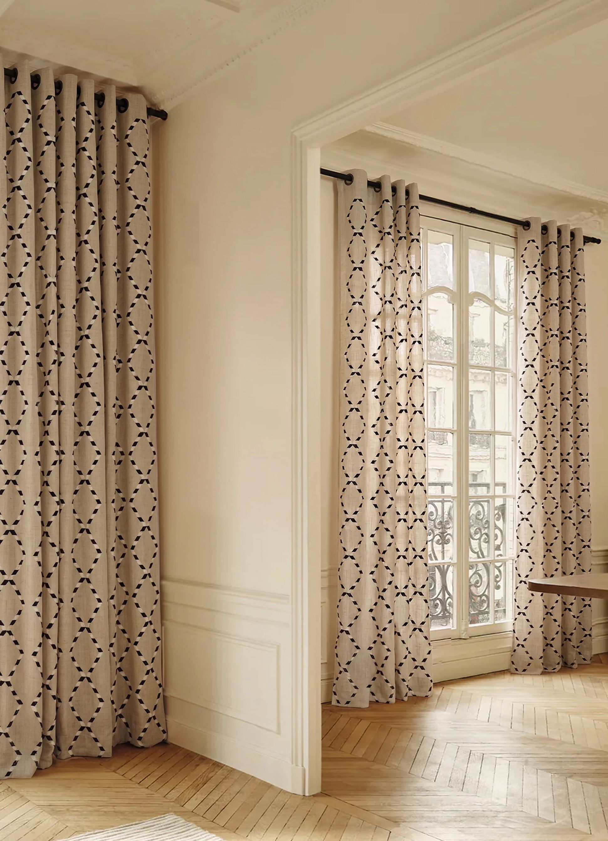 Des rideaux beiges ornés d'un motif géométrique noir sont suspendus dans une pièce élégante dotée d'un parquet clair et d'une grande fenêtre.