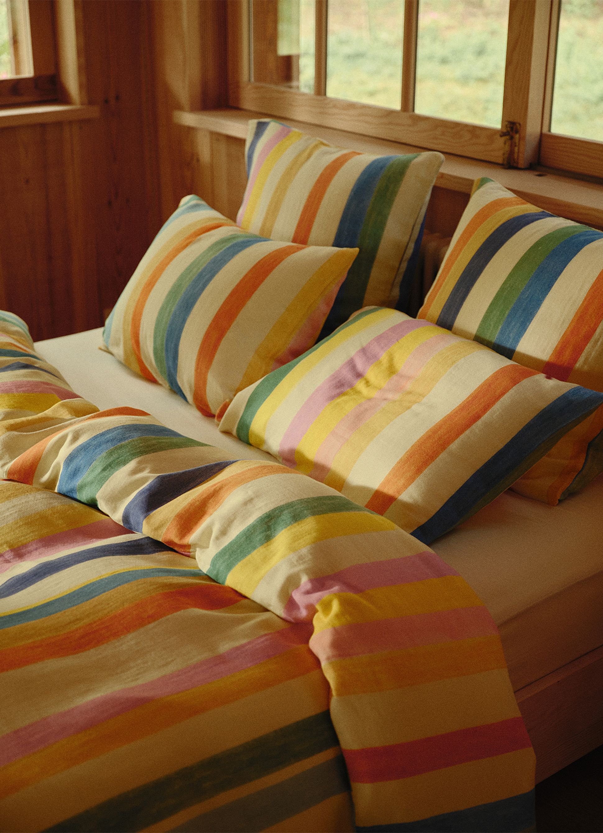 Une parure de lit à rayures colorées, avec des oreillers et une couette, dans une chambre aux boiseries et aux grandes fenêtres.