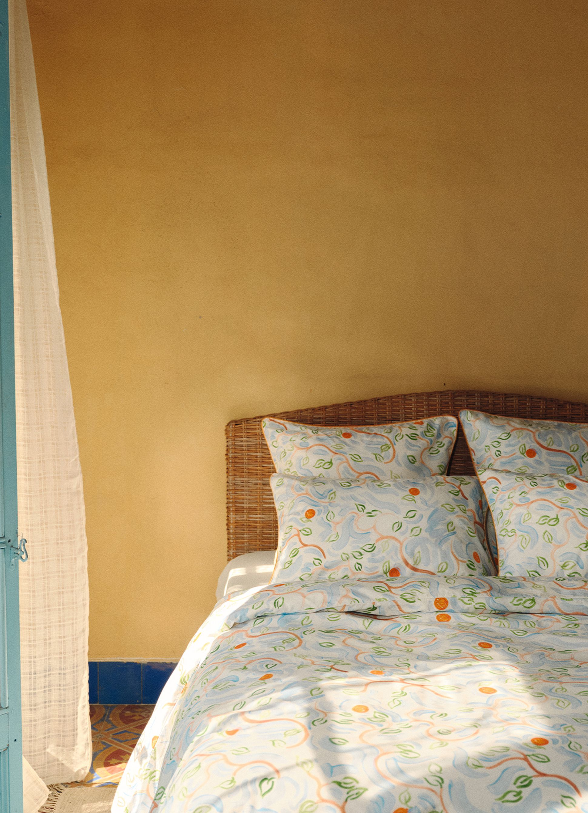 Gros plan sur un lit recouvert de draps colorés à motifs, représentant des cercles orange et des feuilles vertes, partiellement masqués par un rideau léger et vaporeux.