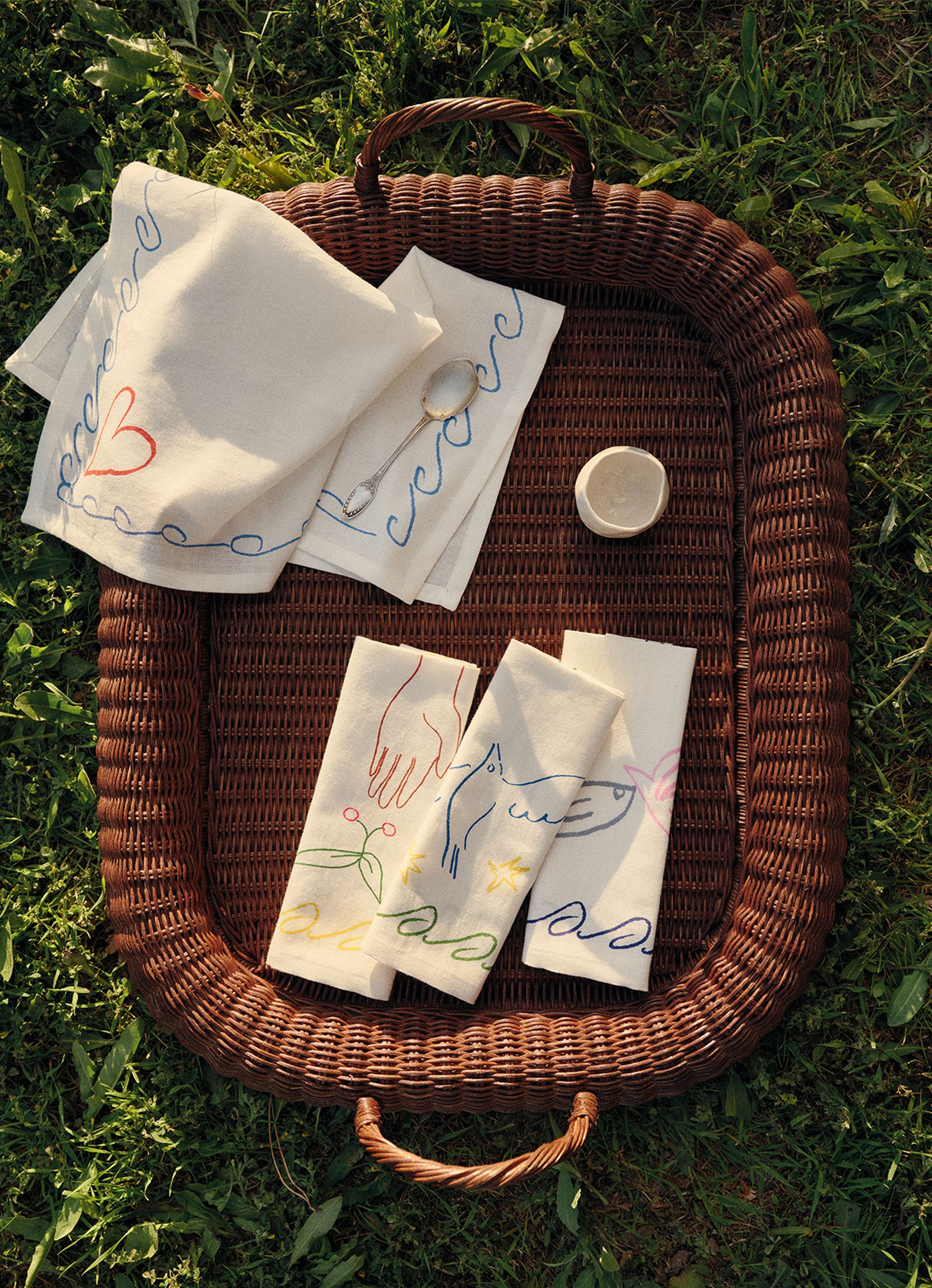 Un plateau en osier posé sur l'herbe contient des serviettes brodées aux motifs colorés, une cuillère et un petit récipient rond.