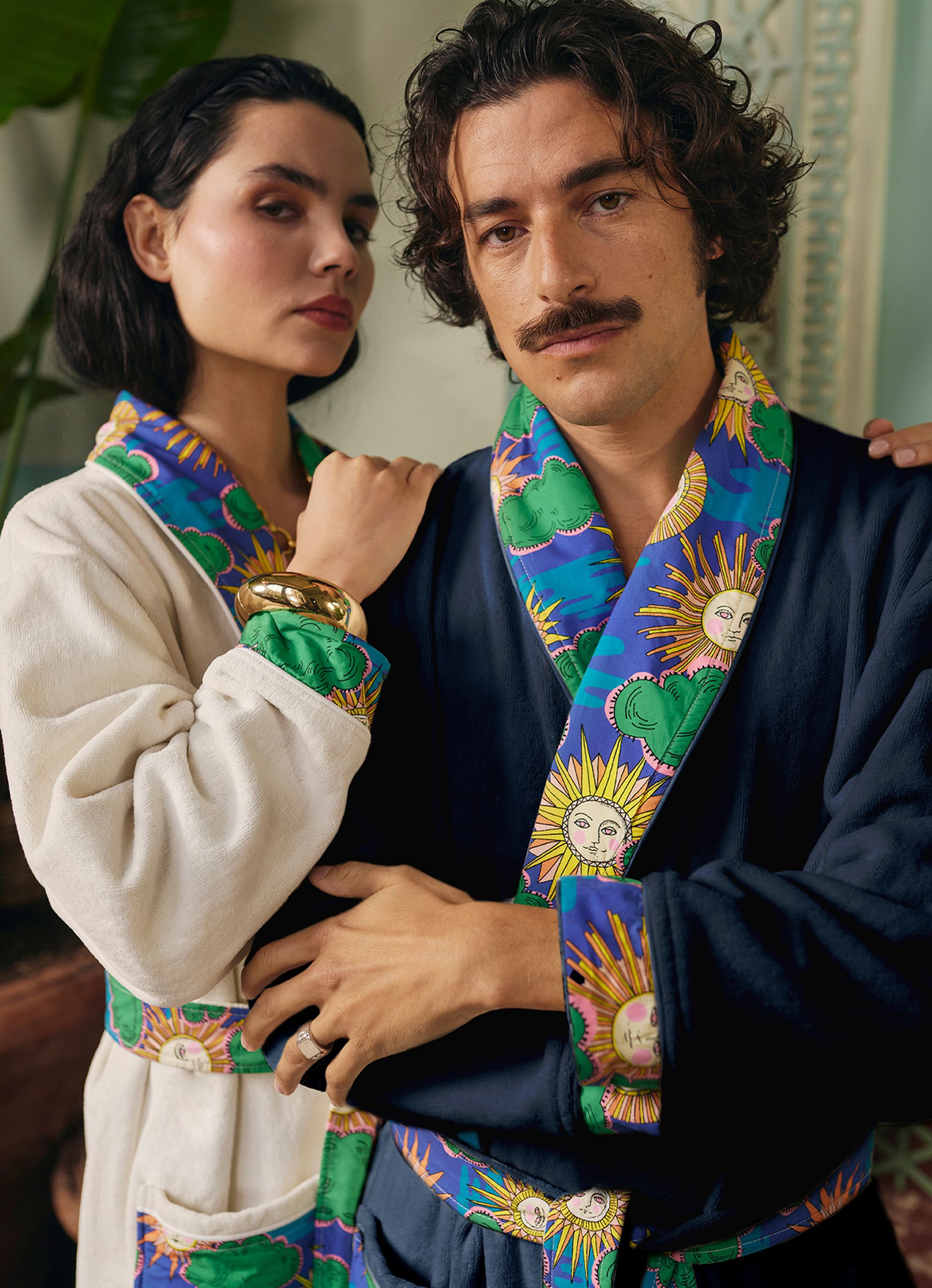 Un homme et une femme vêtus de peignoirs colorés ornés de motifs représentant des soleils et des nuages se tiennent côte à côte, l'air serein et serein, entourés de plantes.
