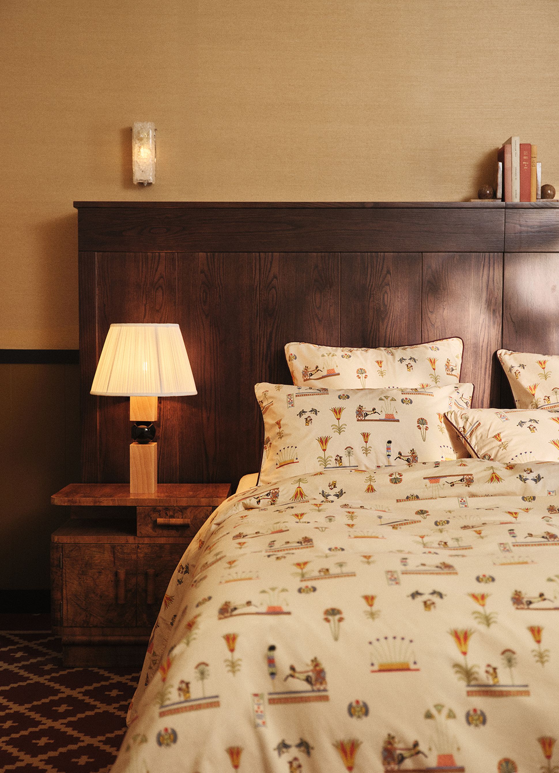Cozy bedroom with a wooden headboard, patterned bedding, a bedside lamp, and a nightstand with books.
