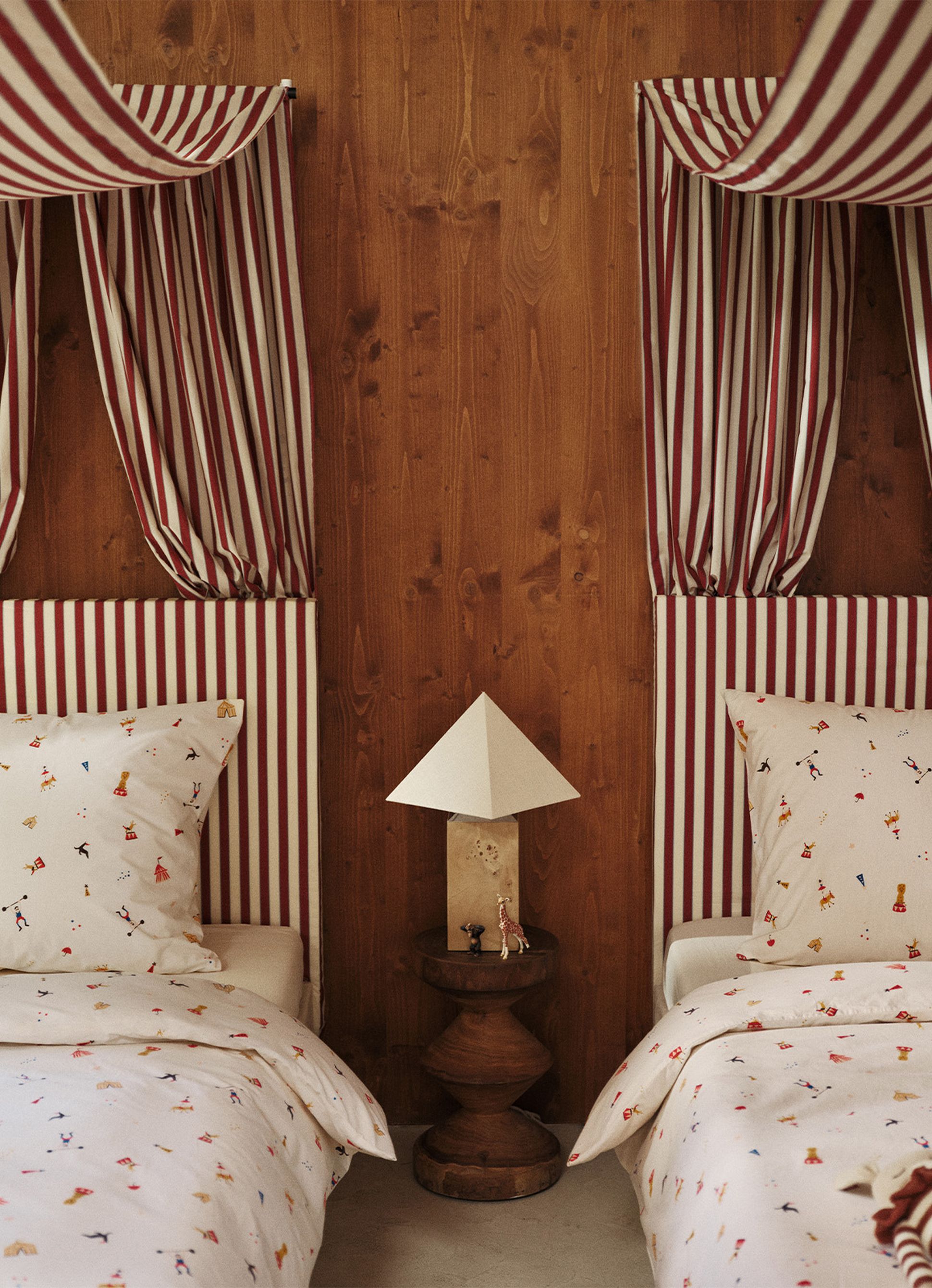 Deux lits jumeaux, dotés de ciels de lit à rayures rouges et d'une literie à motifs, sont séparés par une table de nuit en bois sur laquelle sont posés une lampe en forme de pyramide et de petites figurines.