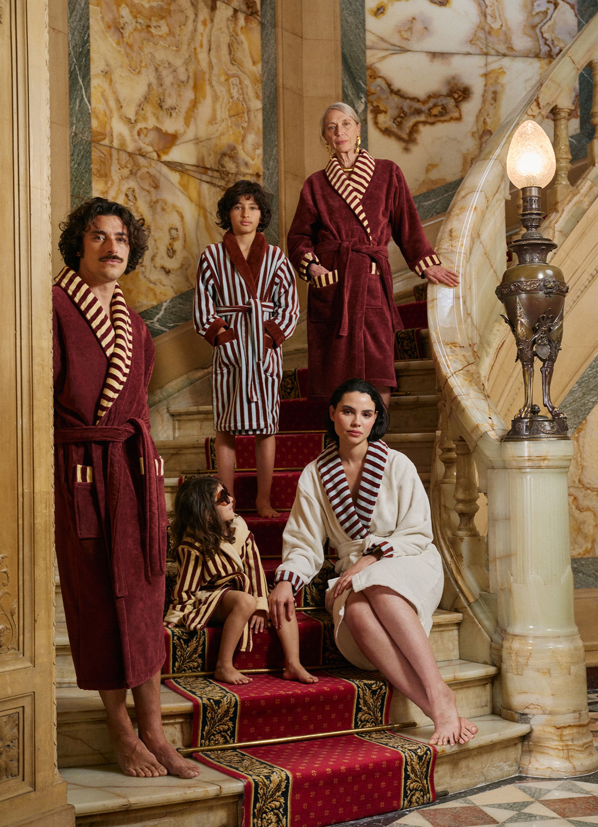 Cinq personnes vêtues de robes somptueuses posent sur un escalier en marbre richement décoré. L'atmosphère est élégante et fastueuse.