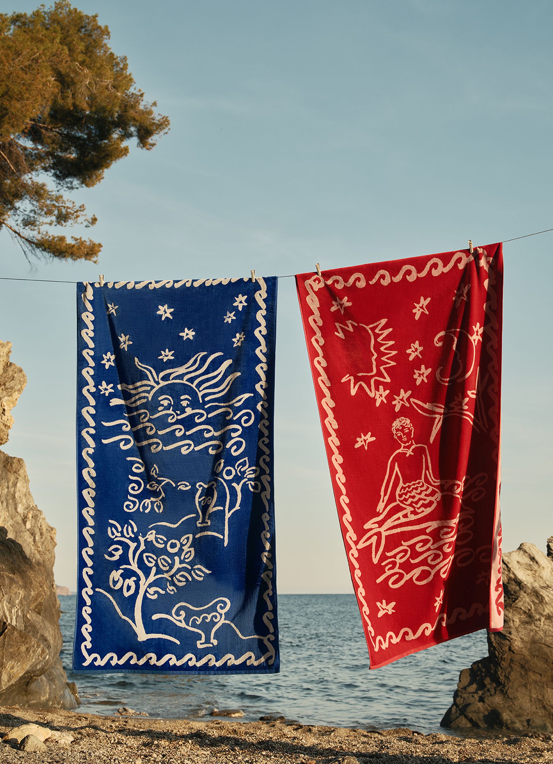 Deux serviettes aux couleurs vives, l'une bleue et l'autre rouge, ornées de motifs artistiques blancs, sont suspendues à une corde près d'une plage rocheuse, sous un ciel dégagé.