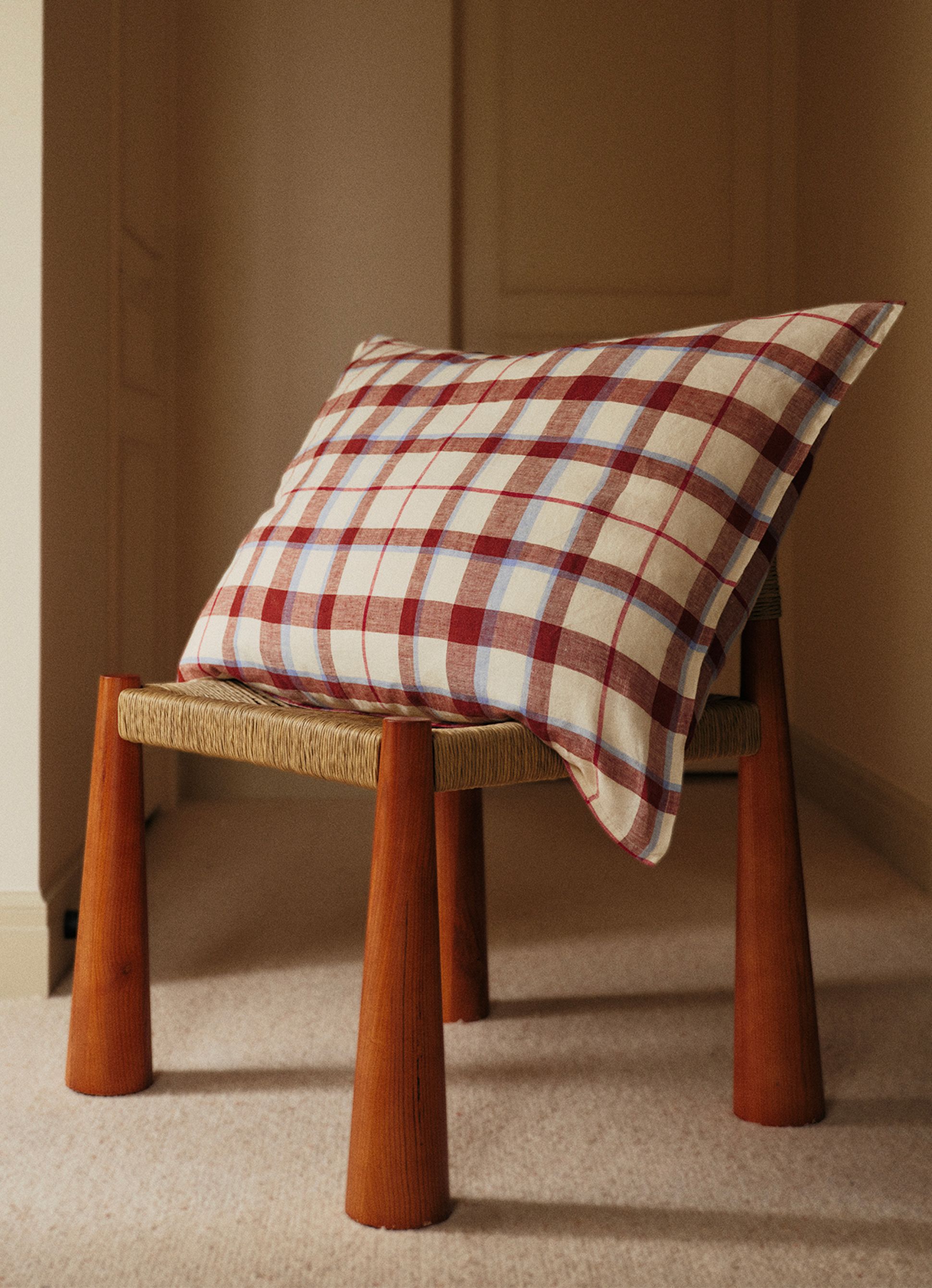Un coussin à carreaux rouges et bleus repose sur un tabouret en bois à assise tressée, posé sur un tapis clair dans une pièce baignée d'une lumière tamisée.