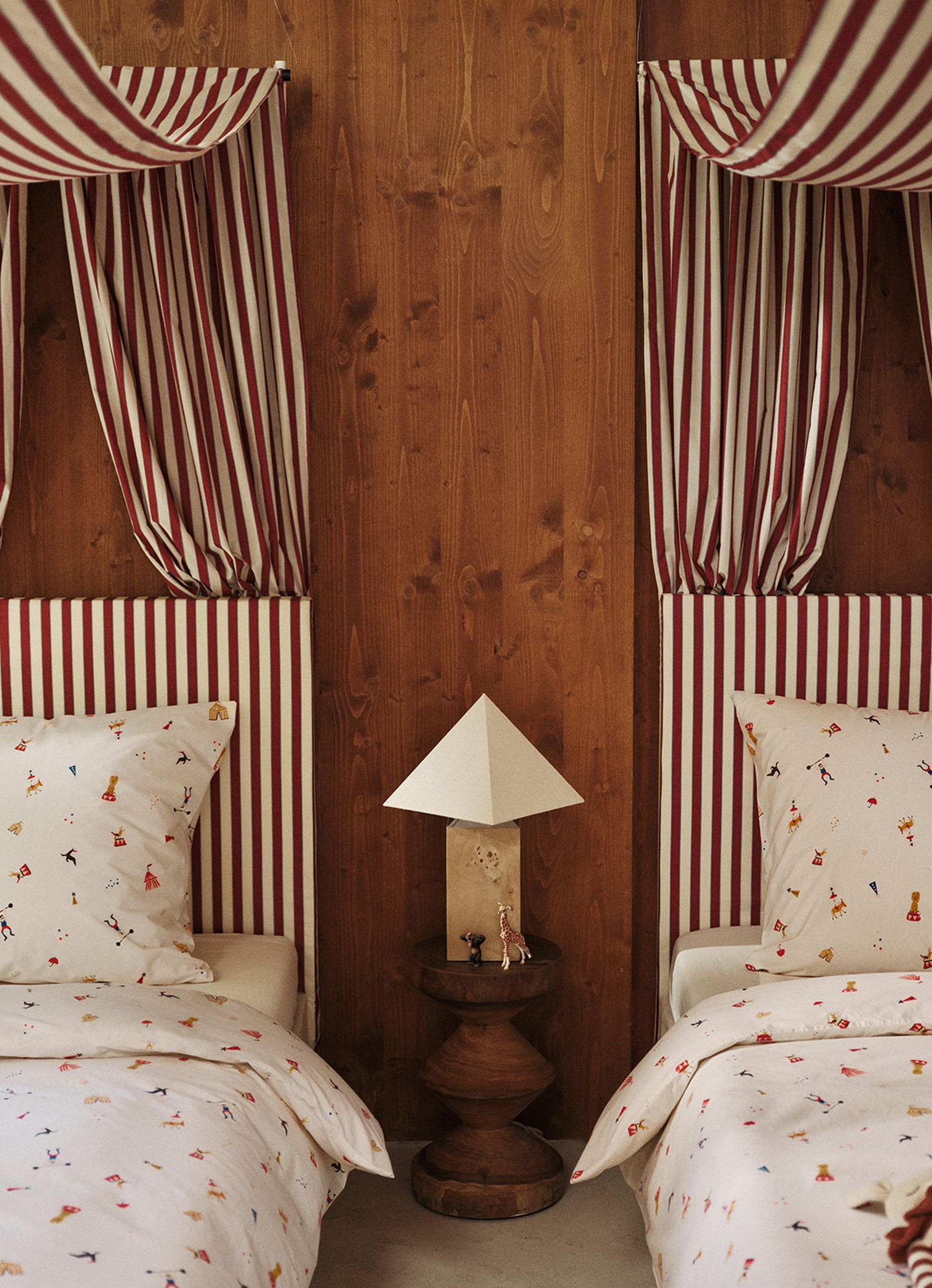 Deux lits avec des baldaquins rayés de rouge et de blanc, une parure de lit blanche aux motifs colorés, ainsi qu'une table de chevet en bois sur laquelle sont posés une lampe et de petites figurines d'animaux.
