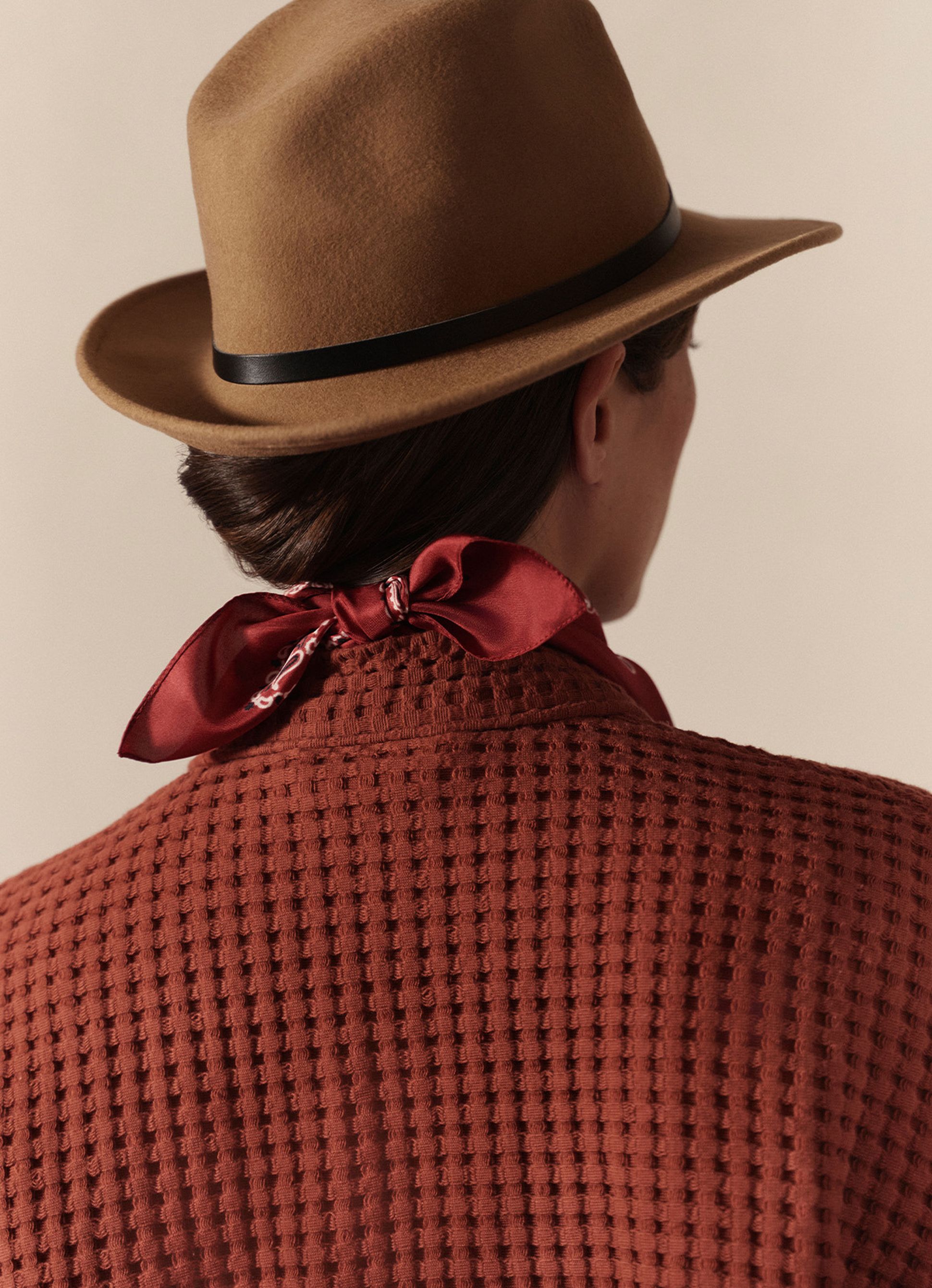 Personne portant un chapeau marron et un peignoir marron texturé, avec une écharpe rouge nouée autour du cou, dos à la caméra, devant un fond neutre.