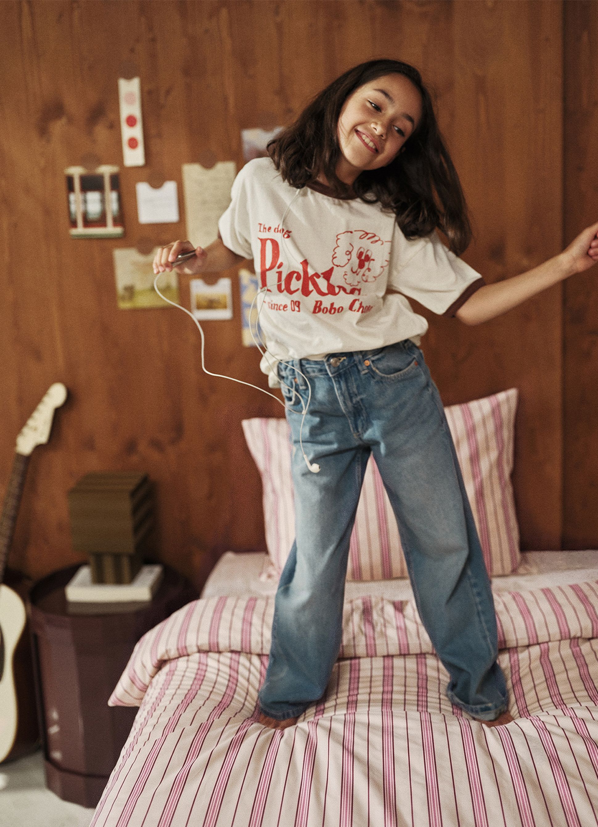 Une jeune fille saute joyeusement sur un lit rayé ; elle porte un casque audio, un t-shirt blanc et un jean, avec une guitare en arrière-plan.