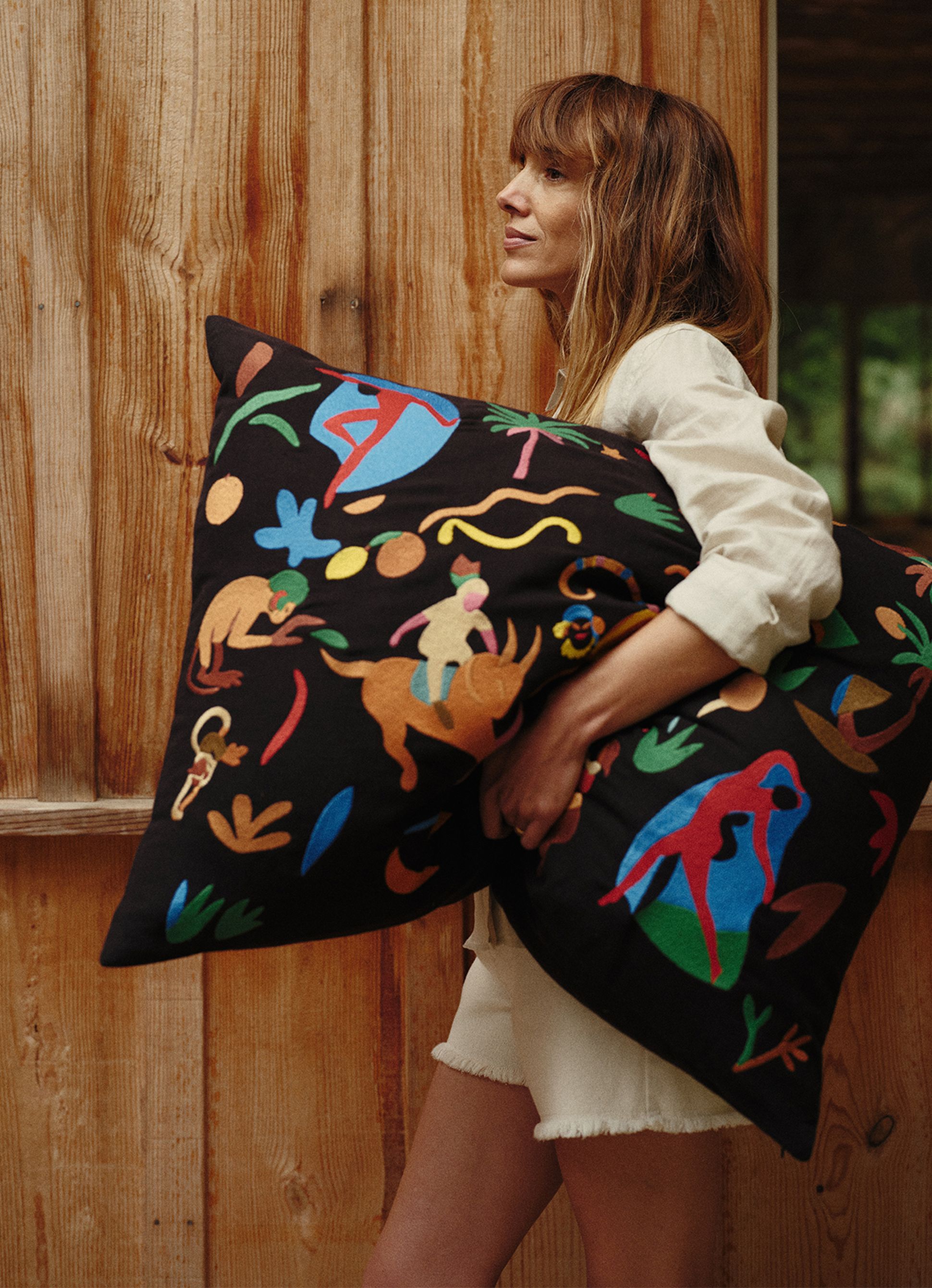Une femme tenant un coussin brodé aux couleurs vives et aux motifs abstraits, debout près d'un mur en bois.